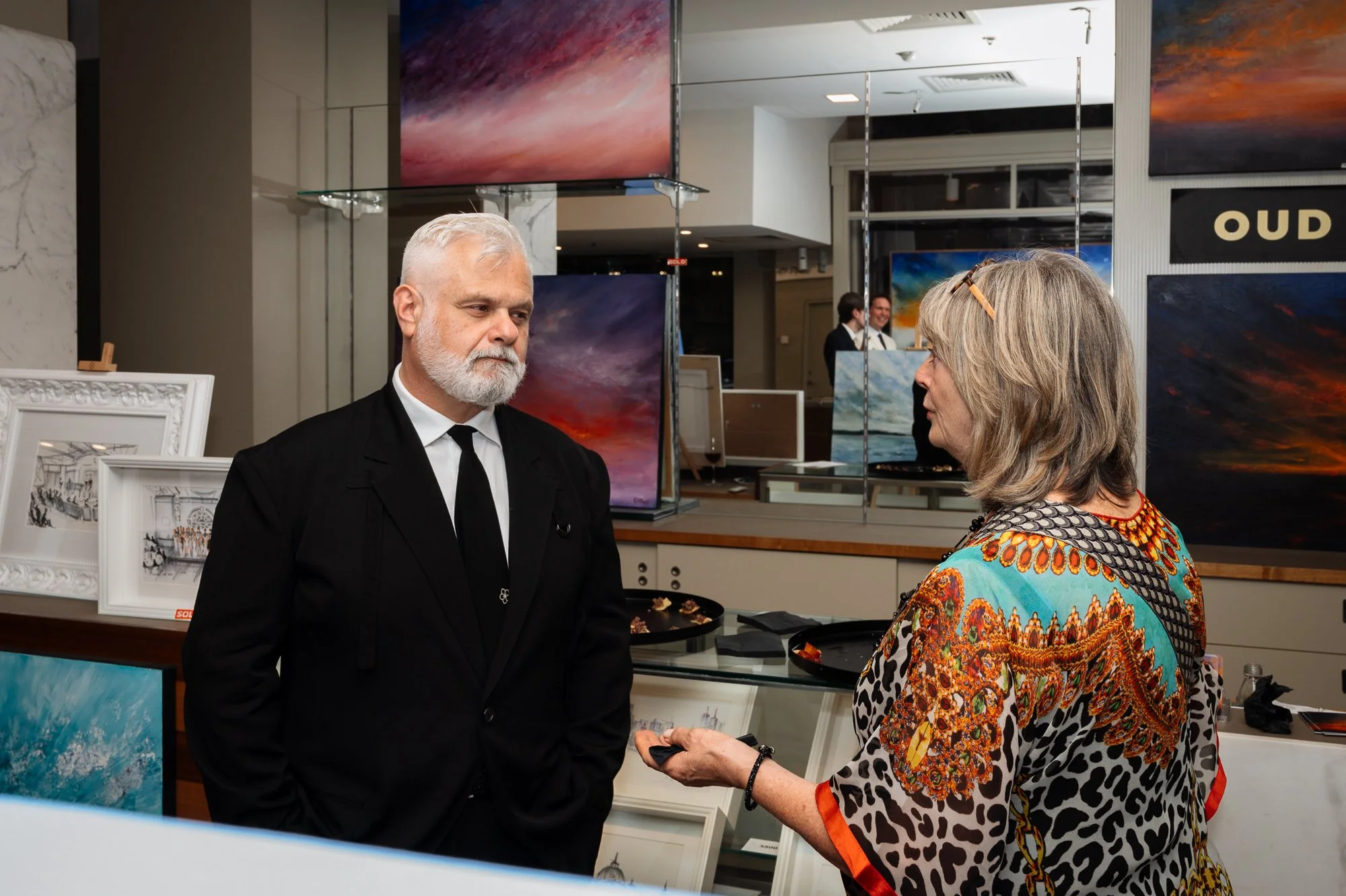 A man with white hair and a beard dressed in a black suit and tie is talking to a woman with shoulder-length gray hair wearing a colorful, patterned blouse at an art gallery or exhibition.