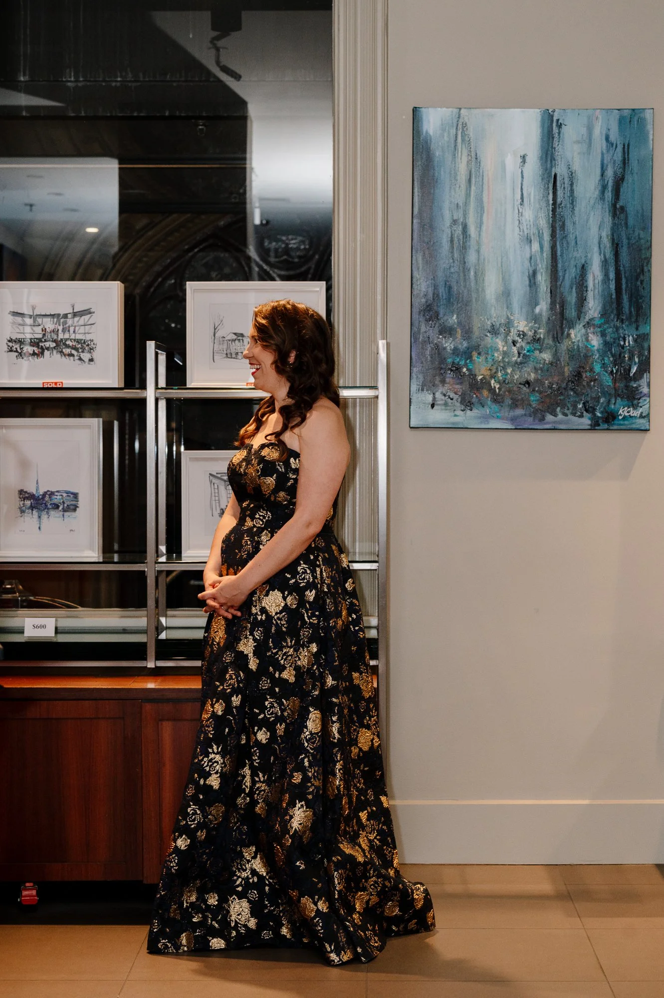 Woman in a black and gold floral dress standing in an art gallery, smiling, with framed artwork behind her and an abstract painting on the wall.