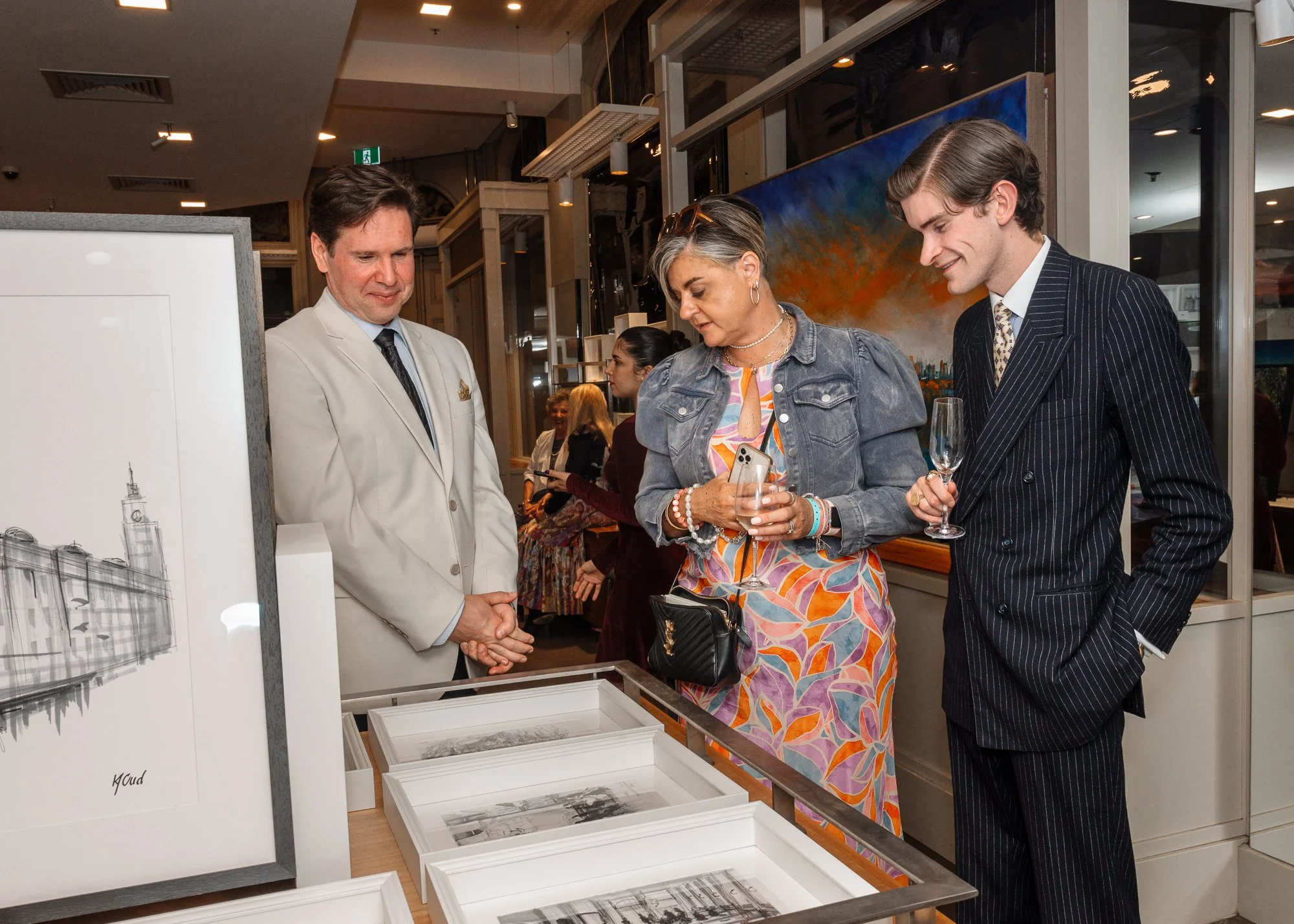 Three people at an art gallery exhibition looking at black and white photographs on display, with one woman holding a glass of champagne and a smartphone, and a man in a pinstripe suit holding a glass of champagne.
