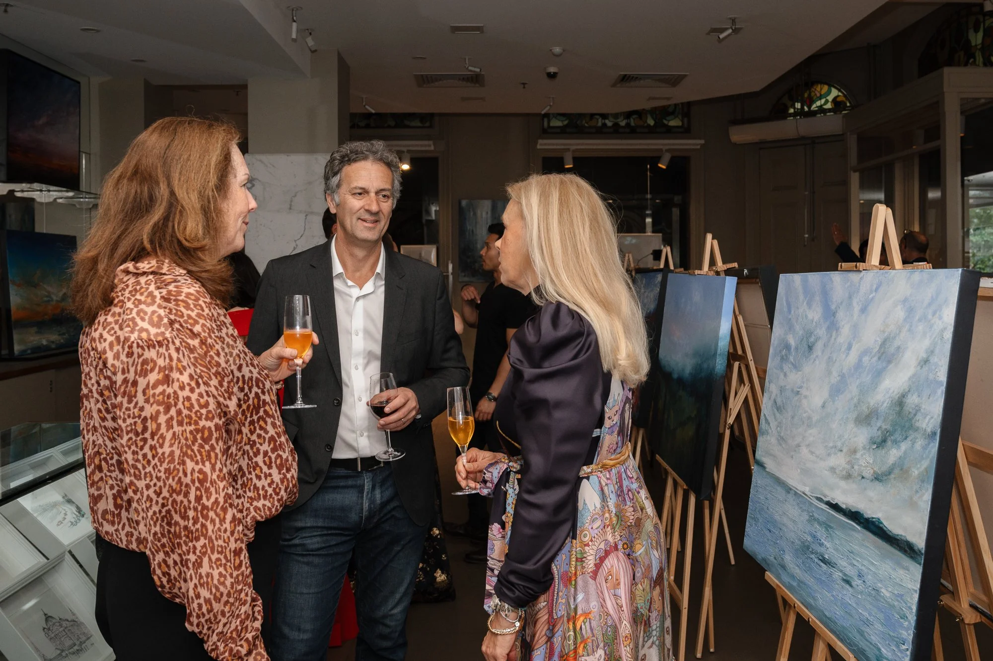 Three people having a conversation at an art exhibit, holding glasses of wine or champagne, with paintings displayed on easels in the background.