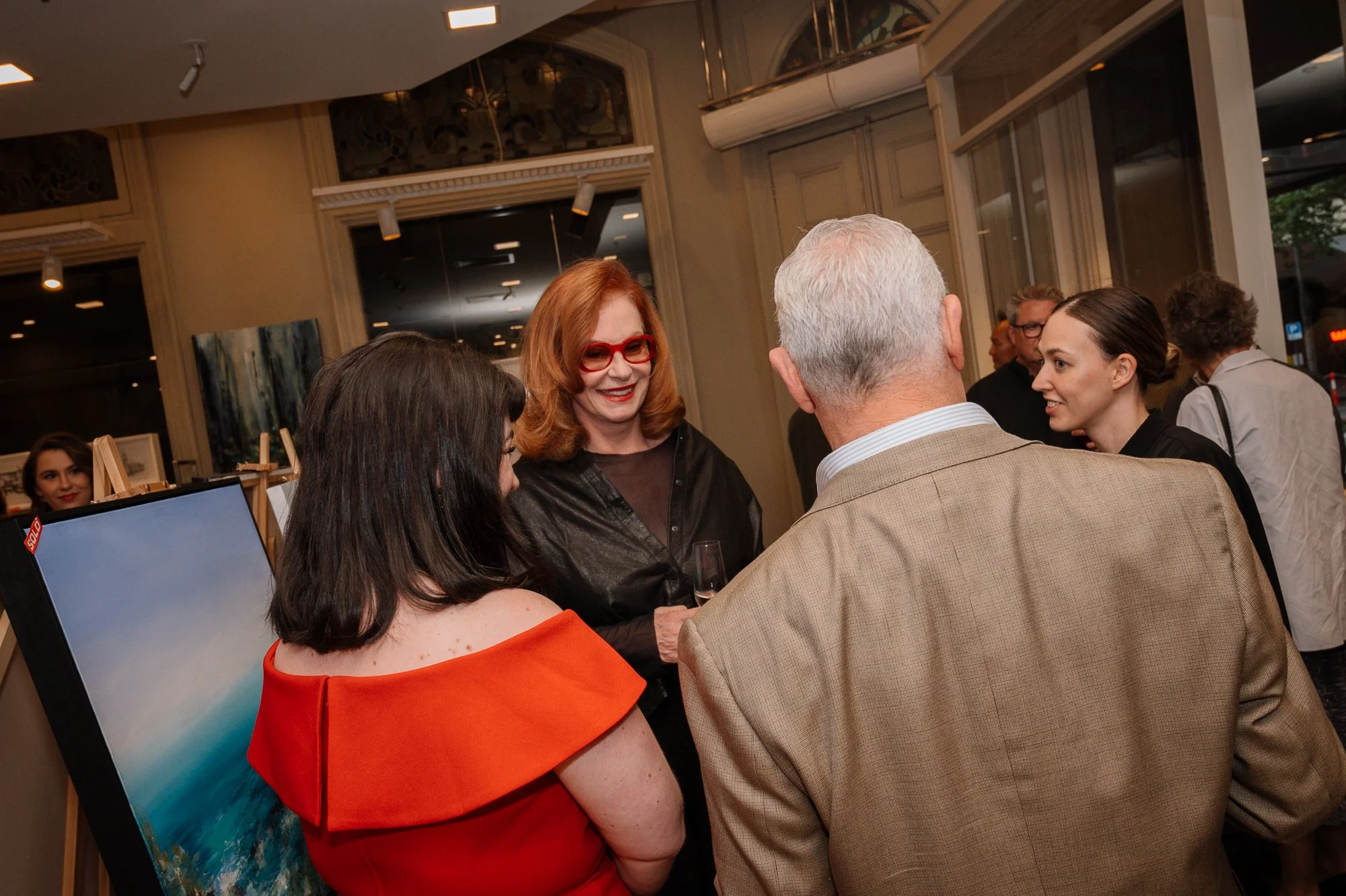 Group of four people talking at an art gallery event, with artwork displayed and other guests in the background.
