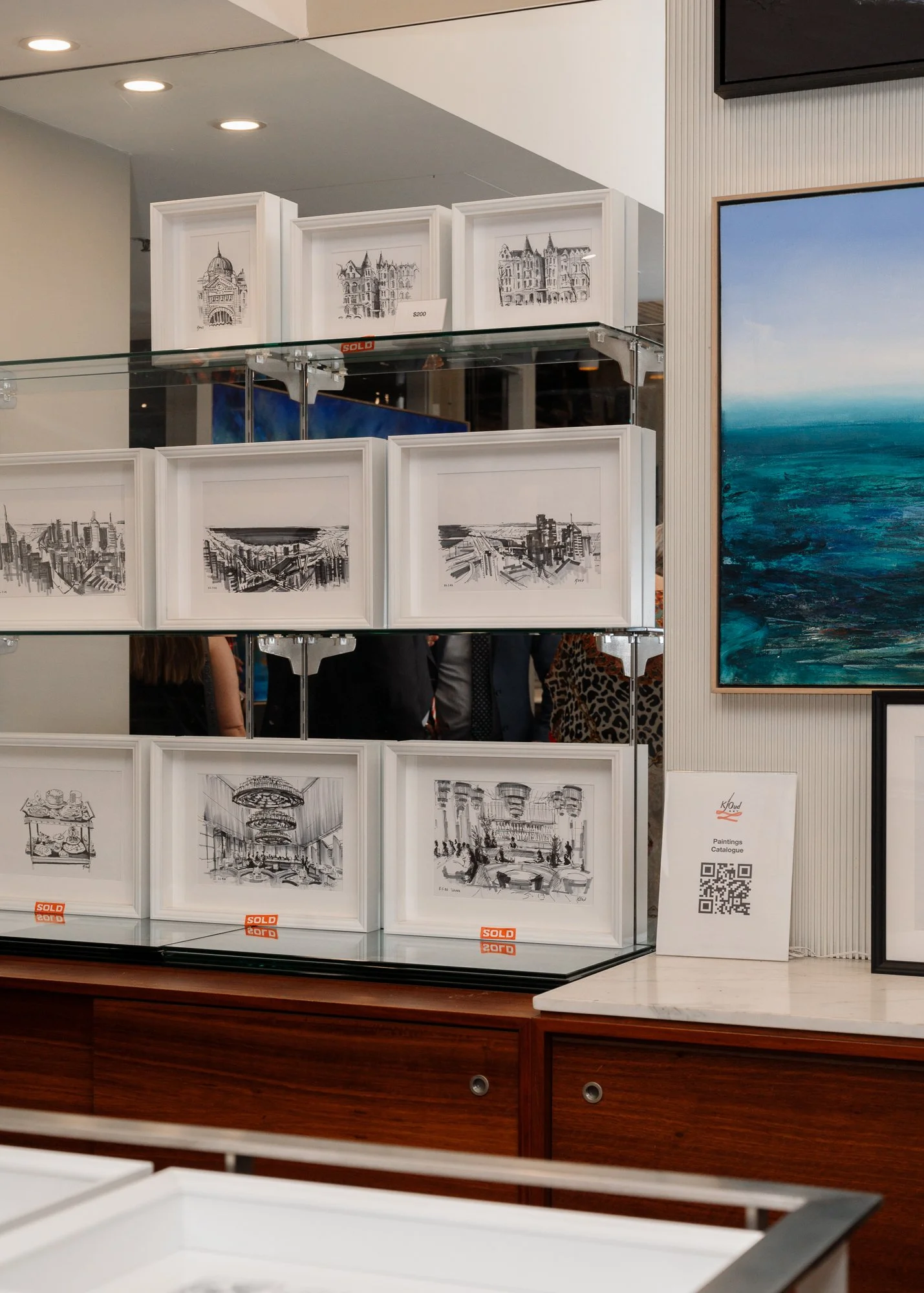 Art gallery display with framed architectural sketches for sale, some marked as sold, next to large landscape painting of ocean.
