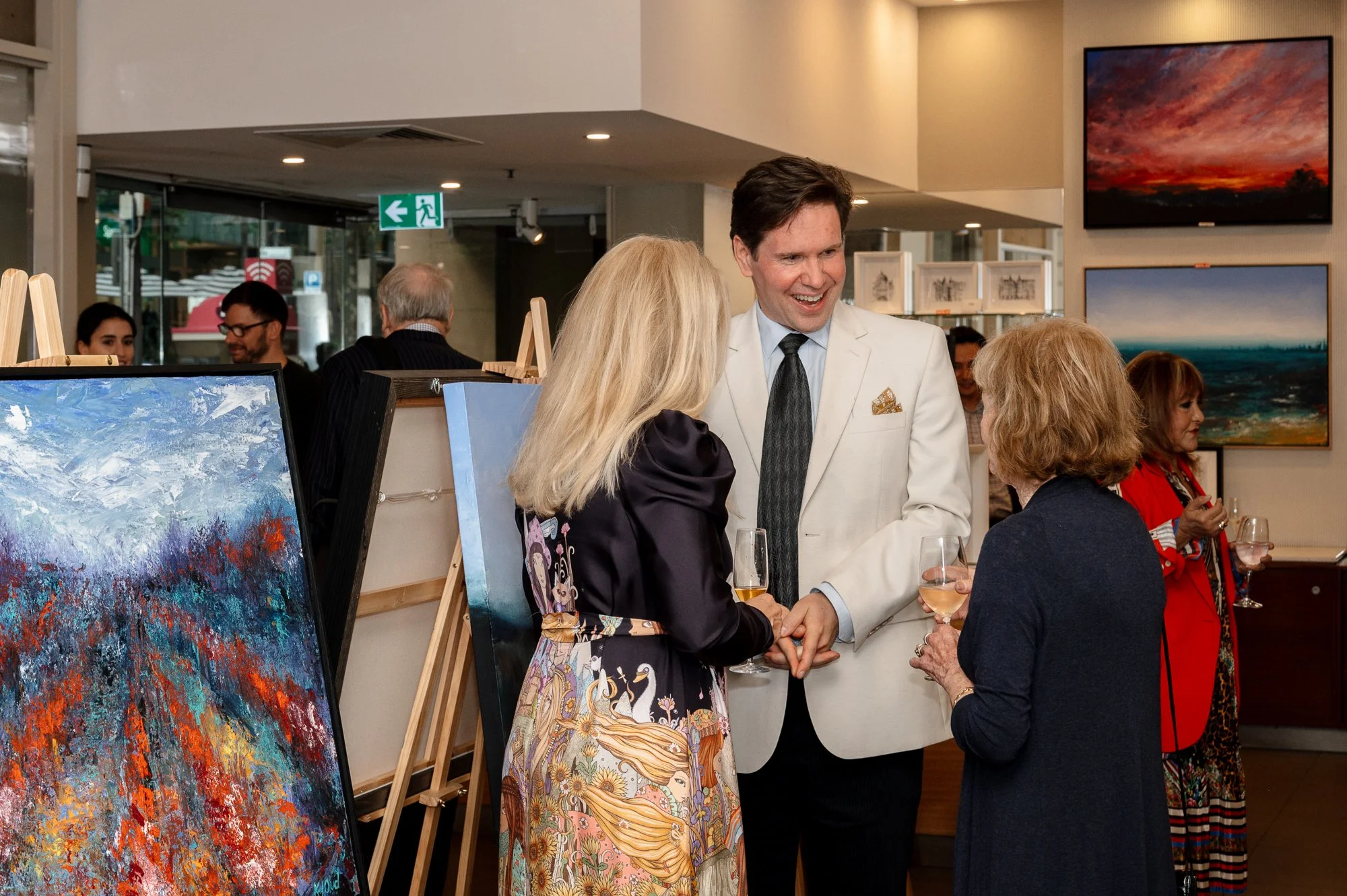People at an art gallery. Three women and one man are talking, holding glasses of wine, with paintings displayed on the walls.