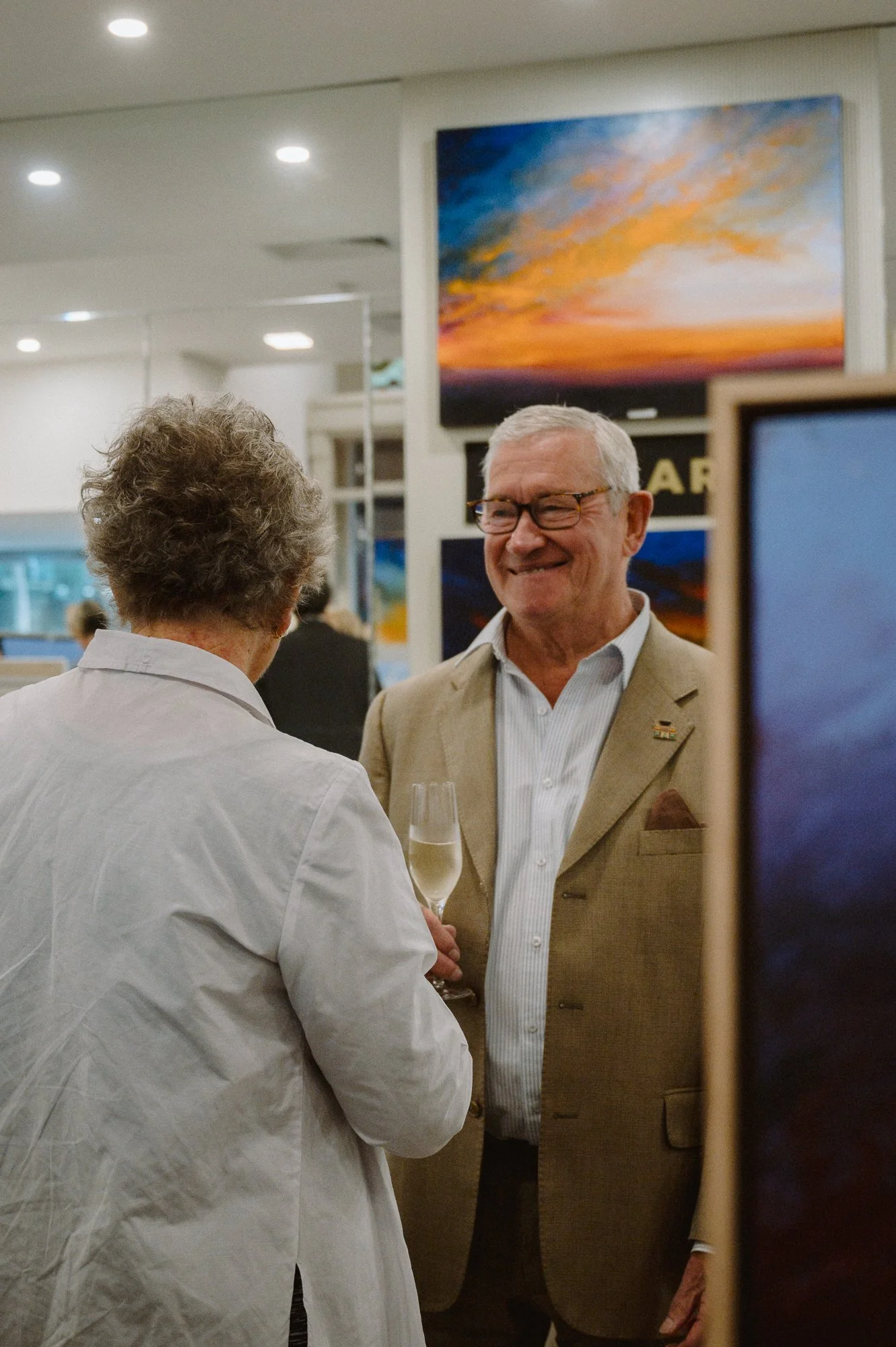 Two people engaged in conversation at an art gallery or exhibition, with paintings on the wall behind them and a television or digital screen visible.