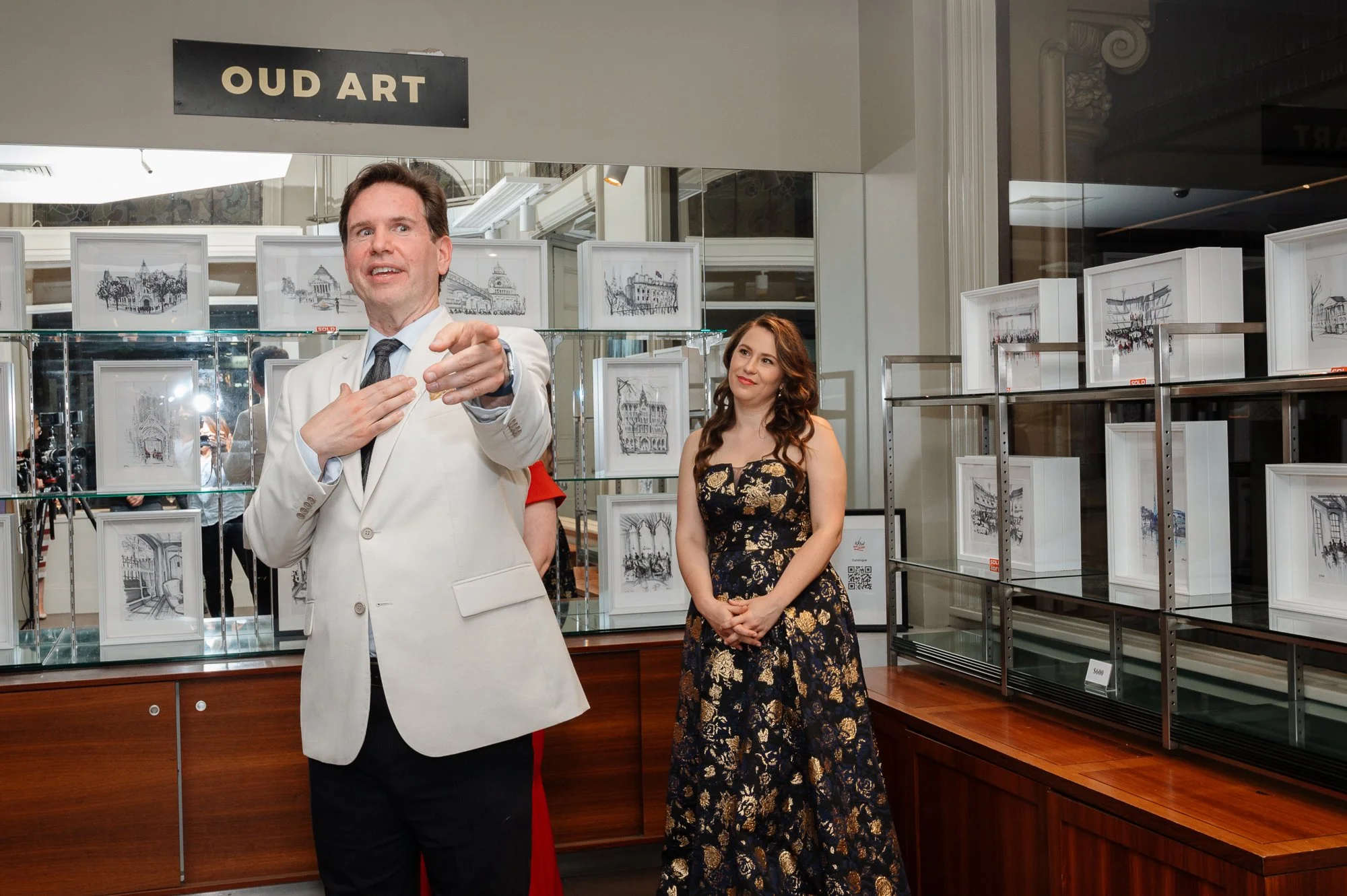 An art exhibit titled 'Oud Art' with framed black-and-white sketches of buildings displayed on glass shelves. A man in a white suit jacket and a woman in a black and gold dress are present, with the man speaking and the woman listening attentively.