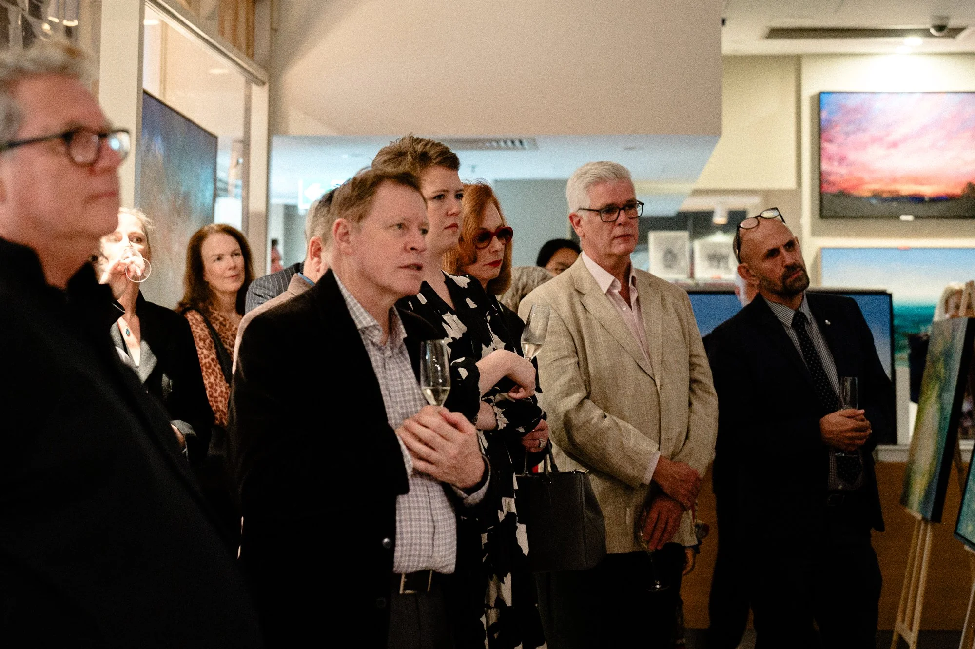 People attending an art exhibition, viewing paintings while holding glasses of champagne.