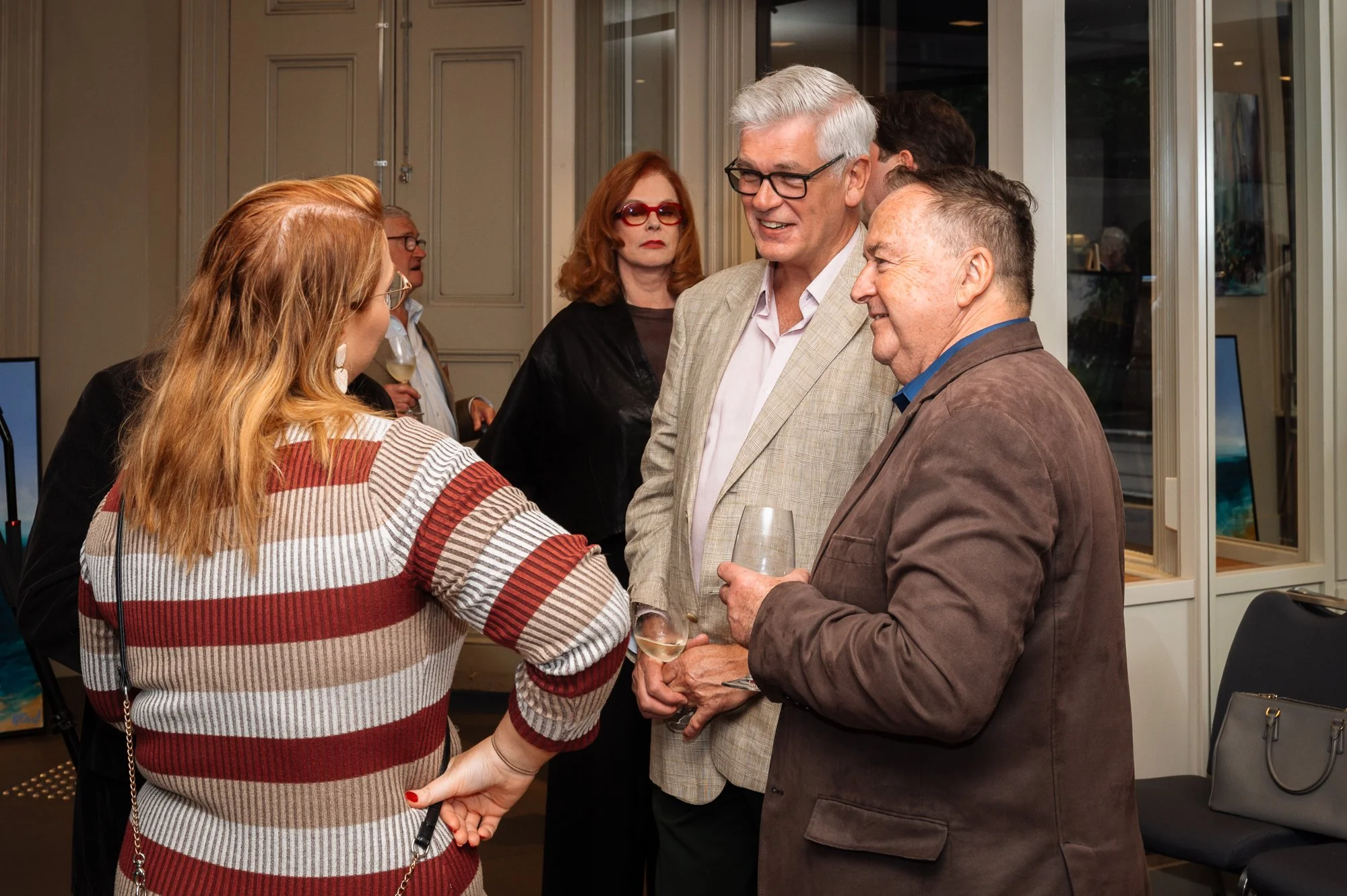 Group of people socializing at a gathering, with three individuals in the foreground engaged in conversation, holding glasses of wine, and others in the background.