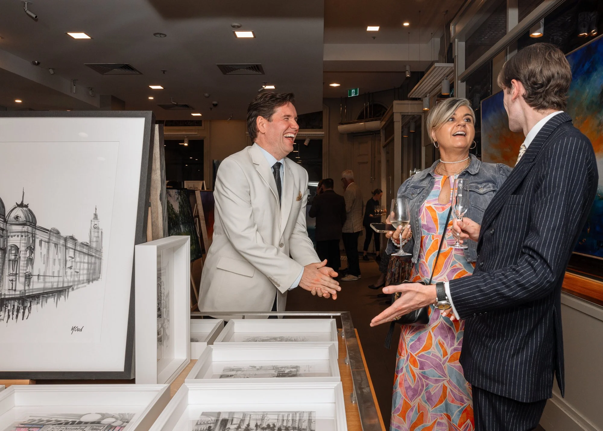 People socializing at an art gallery, with paintings and photos on display, holding drinks and engaging in conversation.