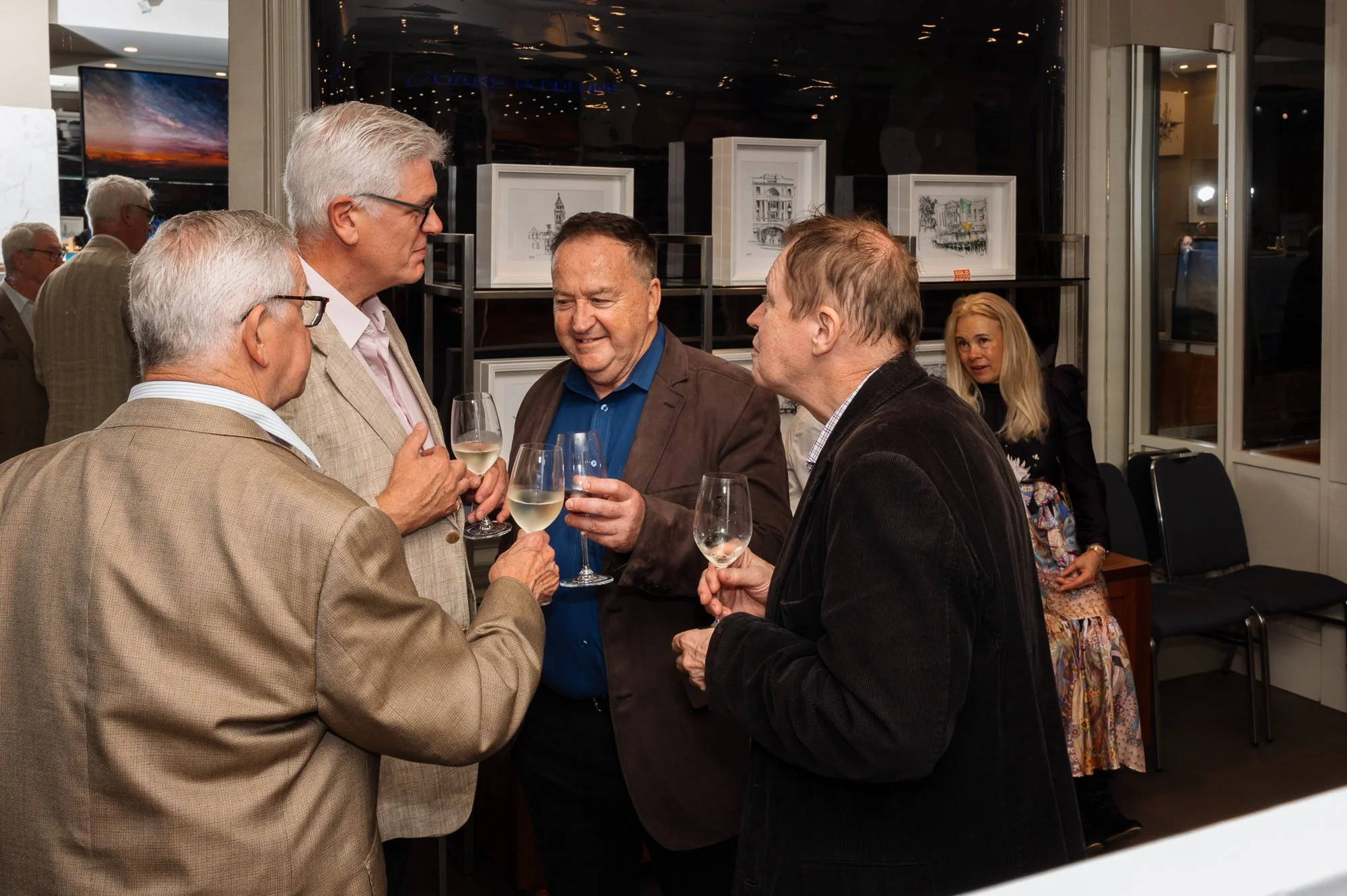 Four middle-aged men in suits are conversing and holding glasses of white wine at a social gathering indoors. A woman with long blonde hair and a patterned dress is seated in the background.