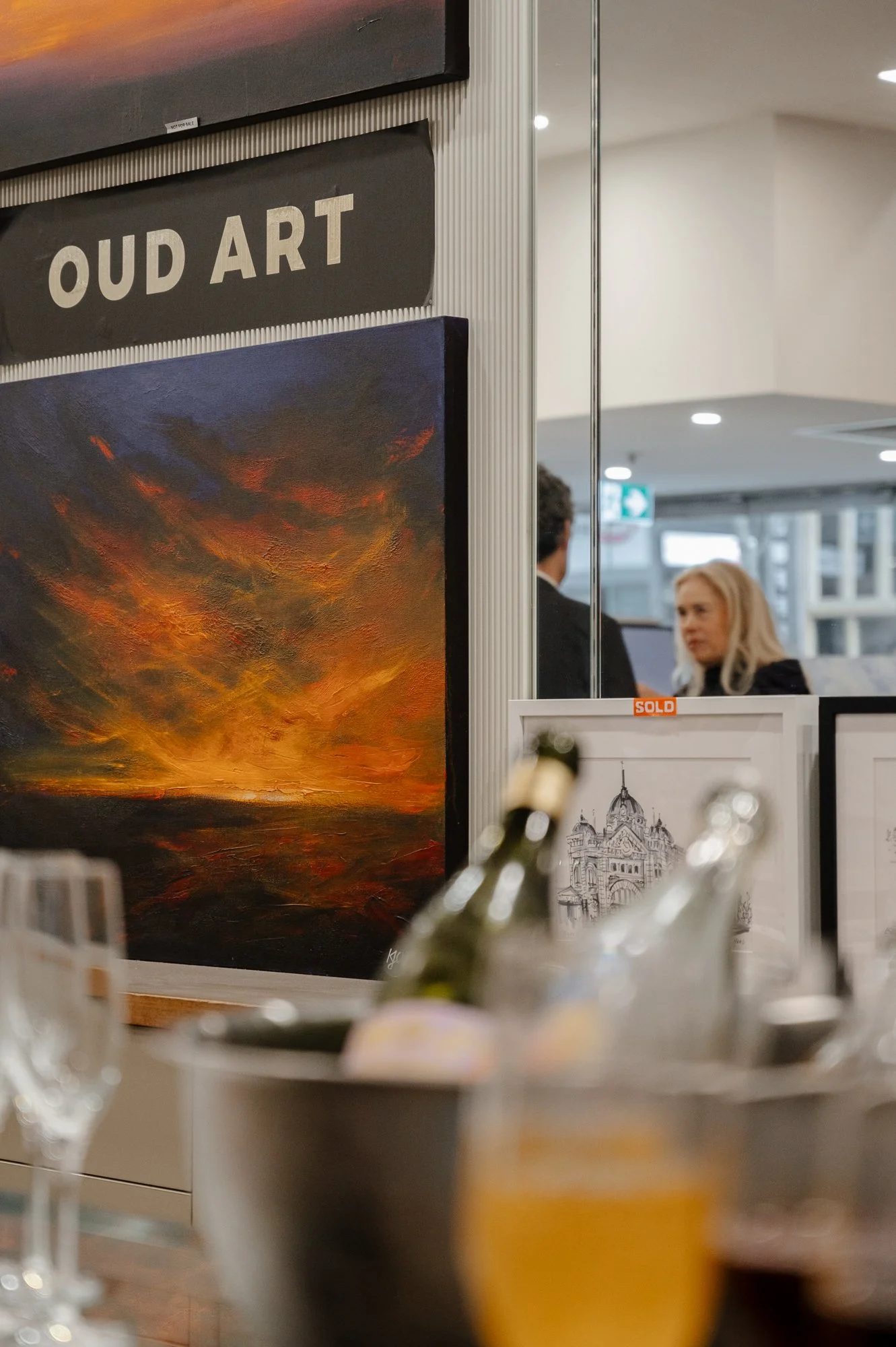 A gallery display with paintings, including a bright sunset or sunrise landscape and a detailed architectural drawing labeled 'sold'. In the background, two women are engaged in conversation, with one facing away and the other looking to her side.