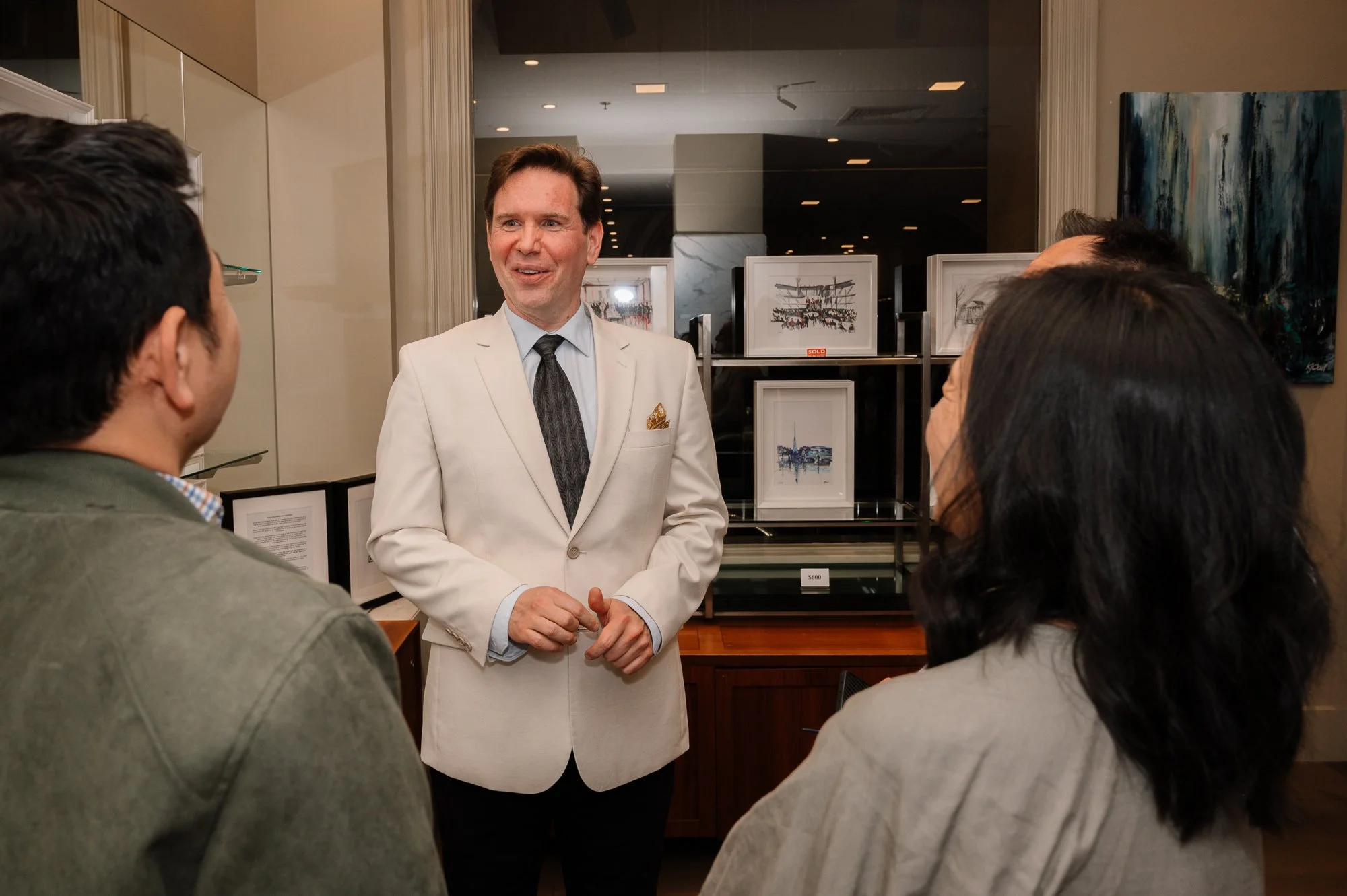 A man in a white blazer and tie speaking with three other people in an art gallery.