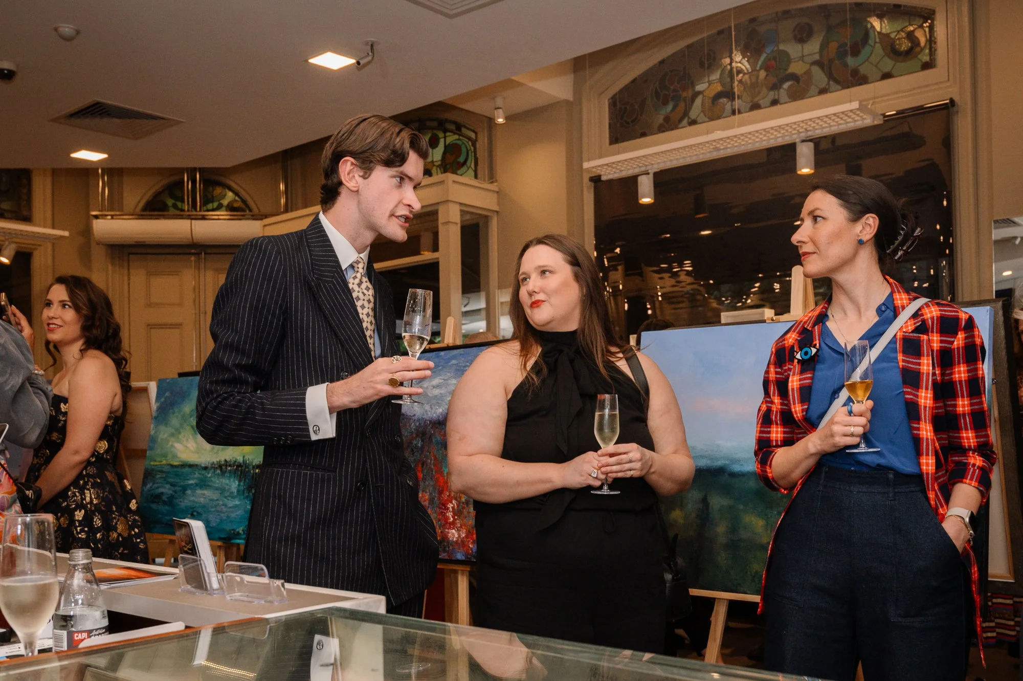 Three people are having a conversation at an art gallery, with paintings displayed behind them. The man on the left has brown hair and is wearing a black pinstripe suit and holding a champagne flute. The woman in the middle has long brown hair and is