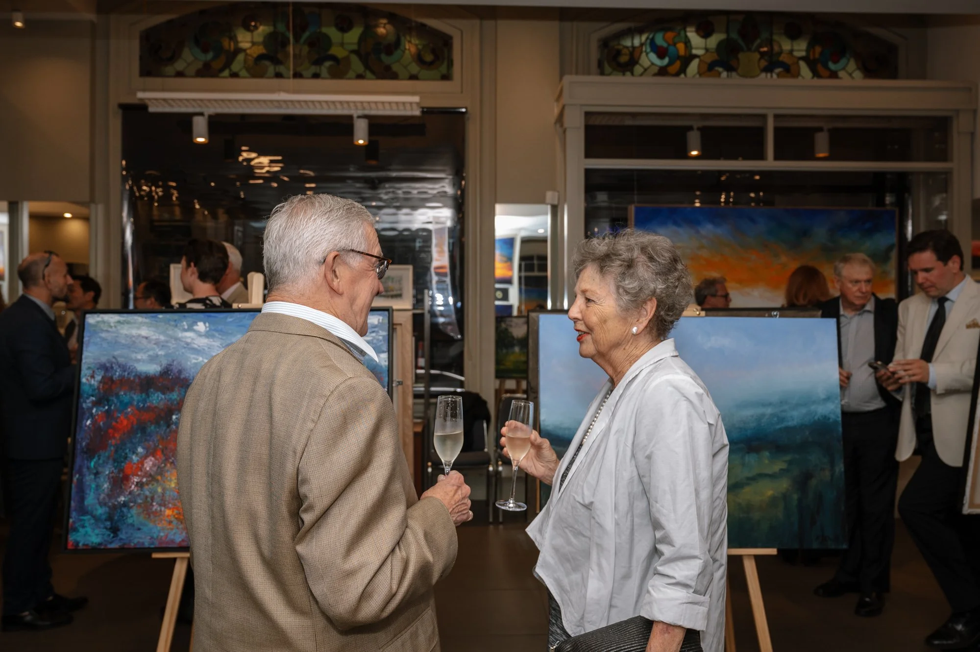 An elderly man and woman talking and holding glasses of champagne at an art gallery, with several paintings on display and other attendees in the background.