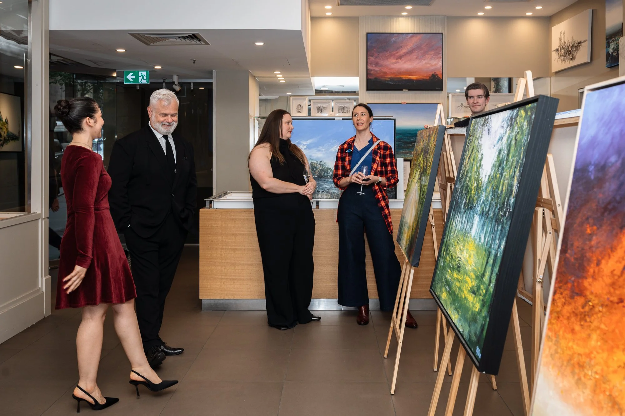 People viewing landscape paintings at an art gallery, with paintings displayed on easels and on the wall in the background.