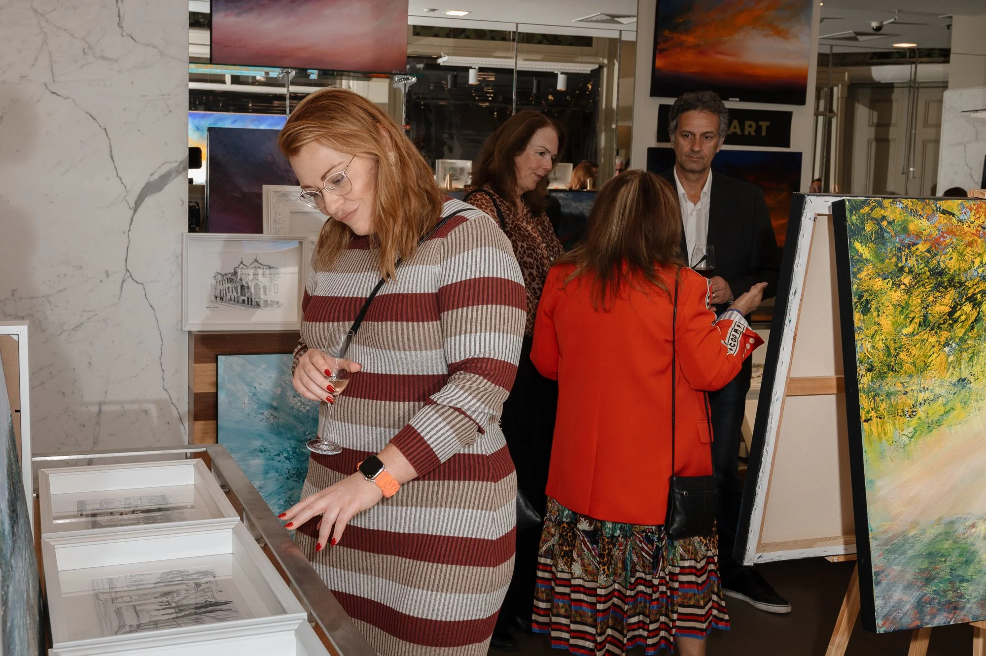 People at an art gallery, viewing paintings and sculptures, with one woman holding a glass of wine.