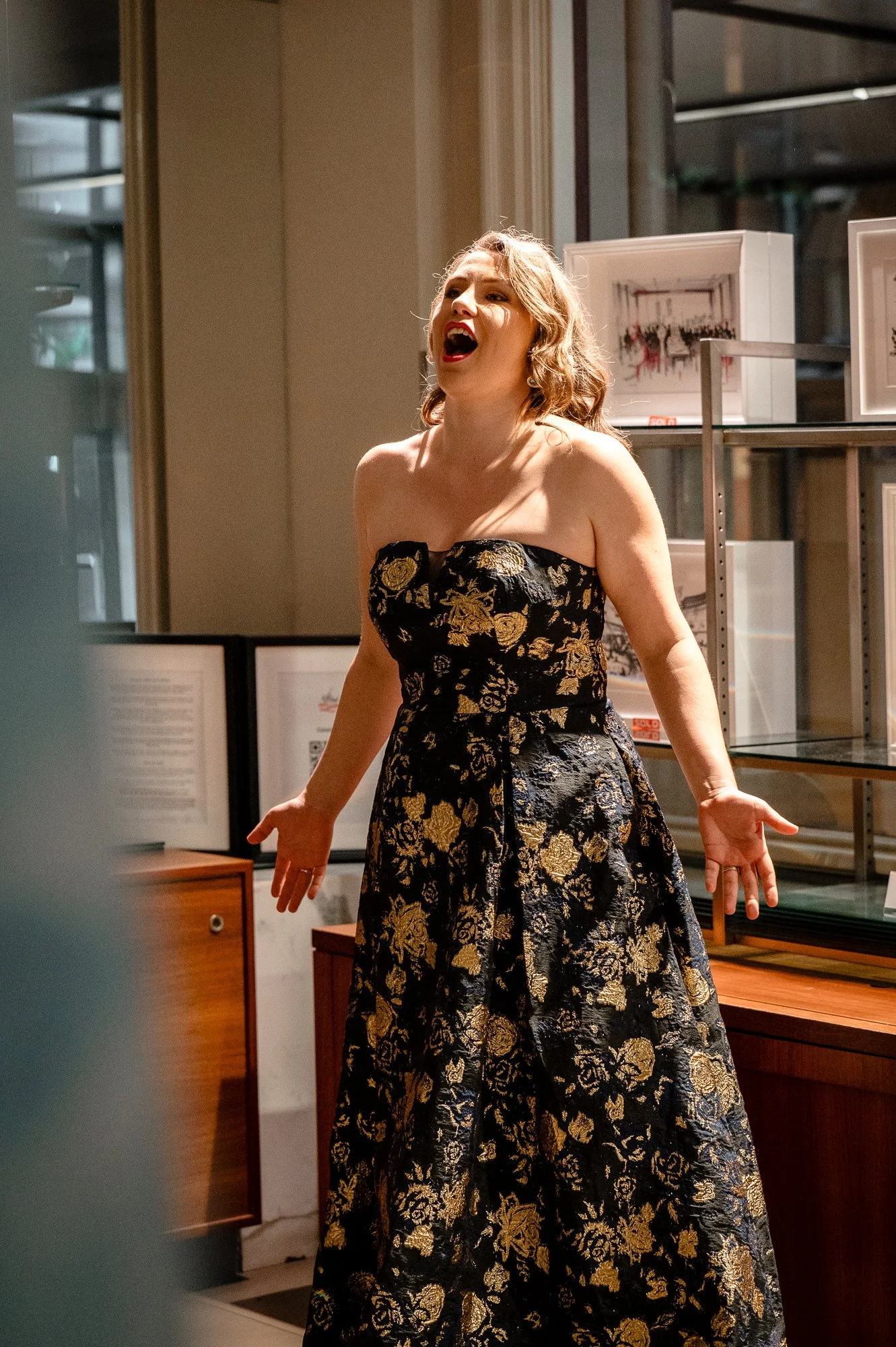 A woman in an elegant black and gold floral strapless dress singing passionately, standing indoors with framed artwork on shelves behind her.