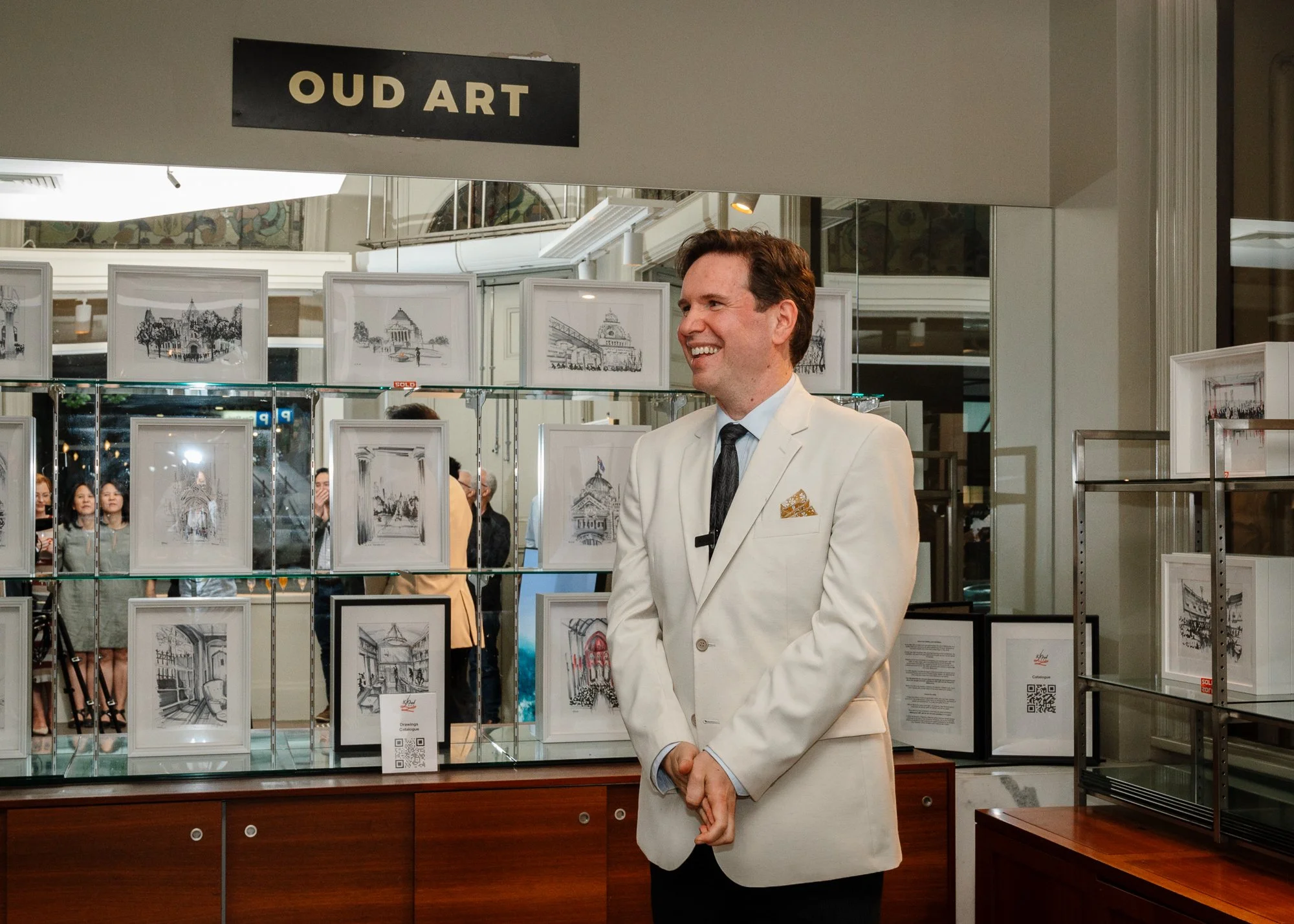 Kristan OUD dressed in a white suit and black tie standing in front of framed artwork at an art exhibition. The sign above reads 'OUD ART' and there are multiple people observing the art in the background.