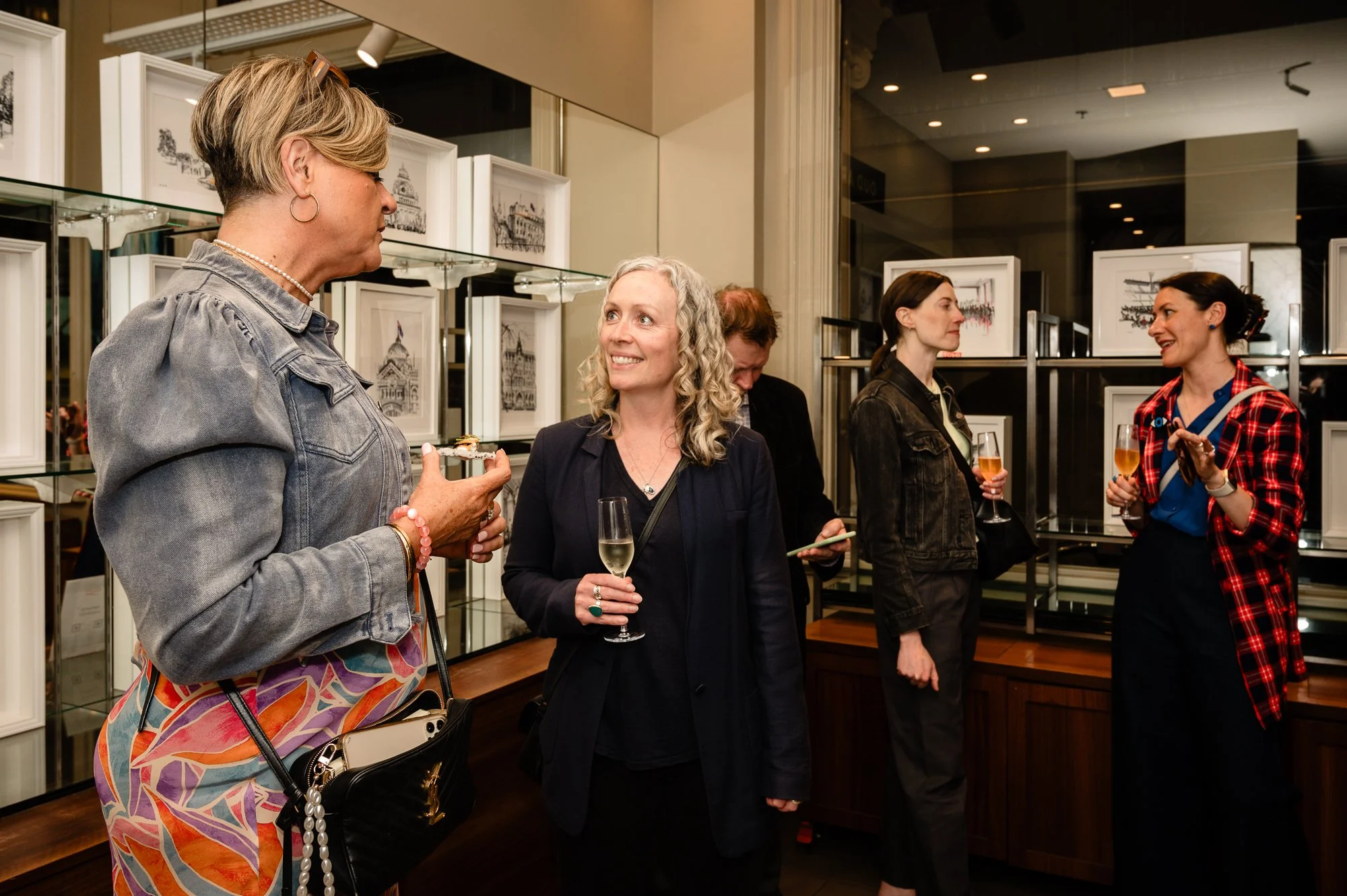 Group of women talking at an art gallery, holding wine glasses, surrounded by interior art displays.
