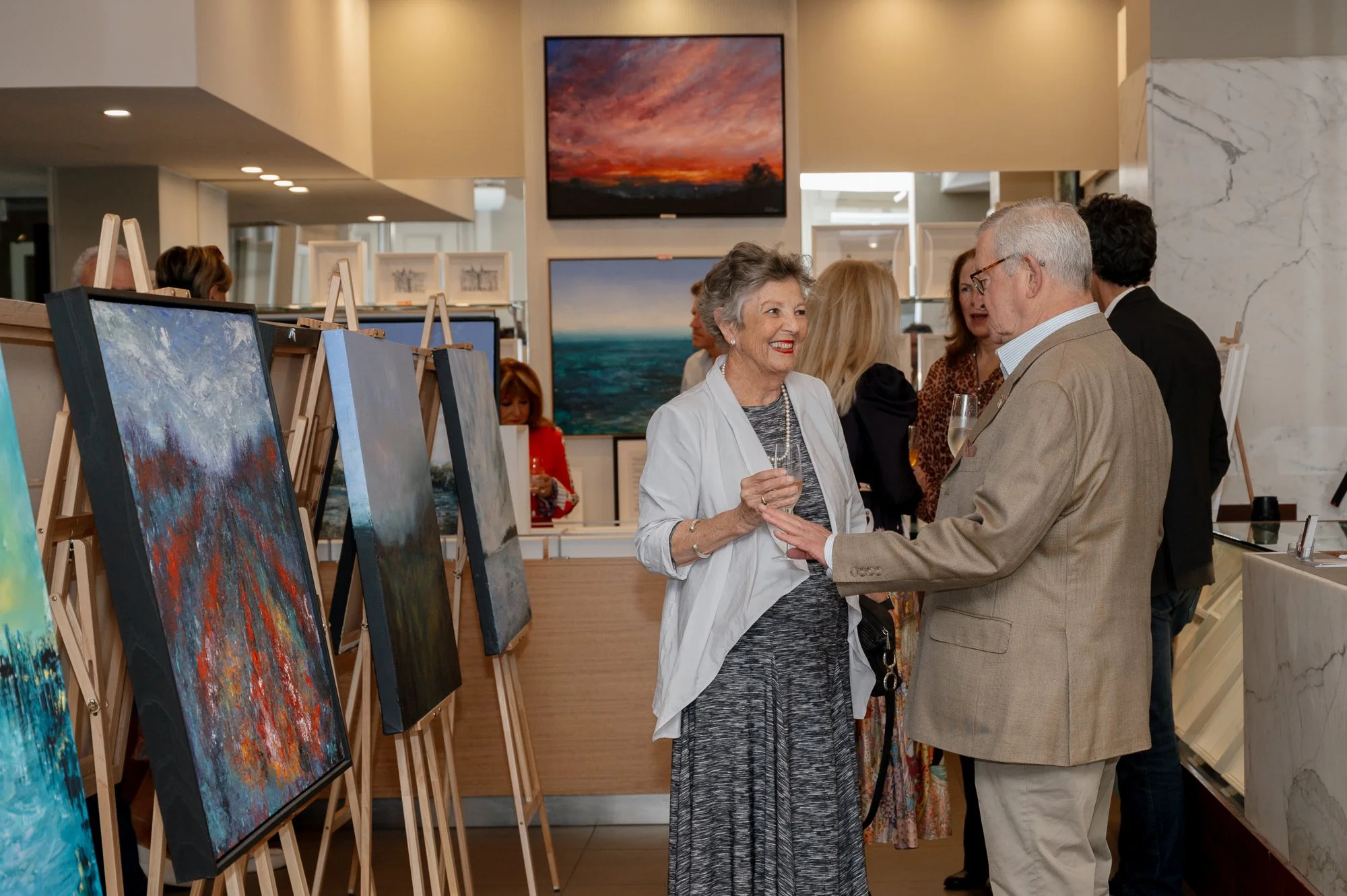 People socializing at an art gallery, with paintings on display along the wall and a couple engaged in conversation in the foreground.