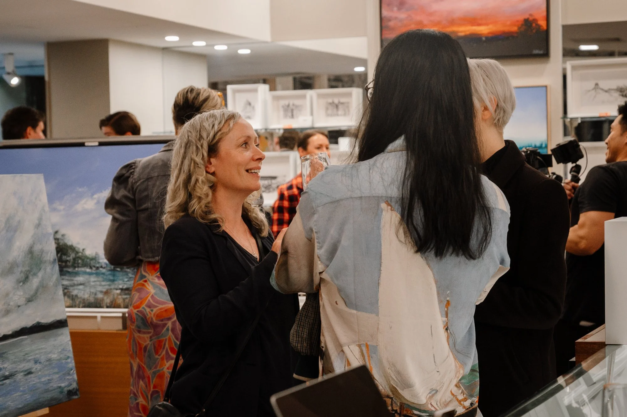 A group of people attending an art exhibition, including women engaging in conversation with paintings and artwork visible in the background.