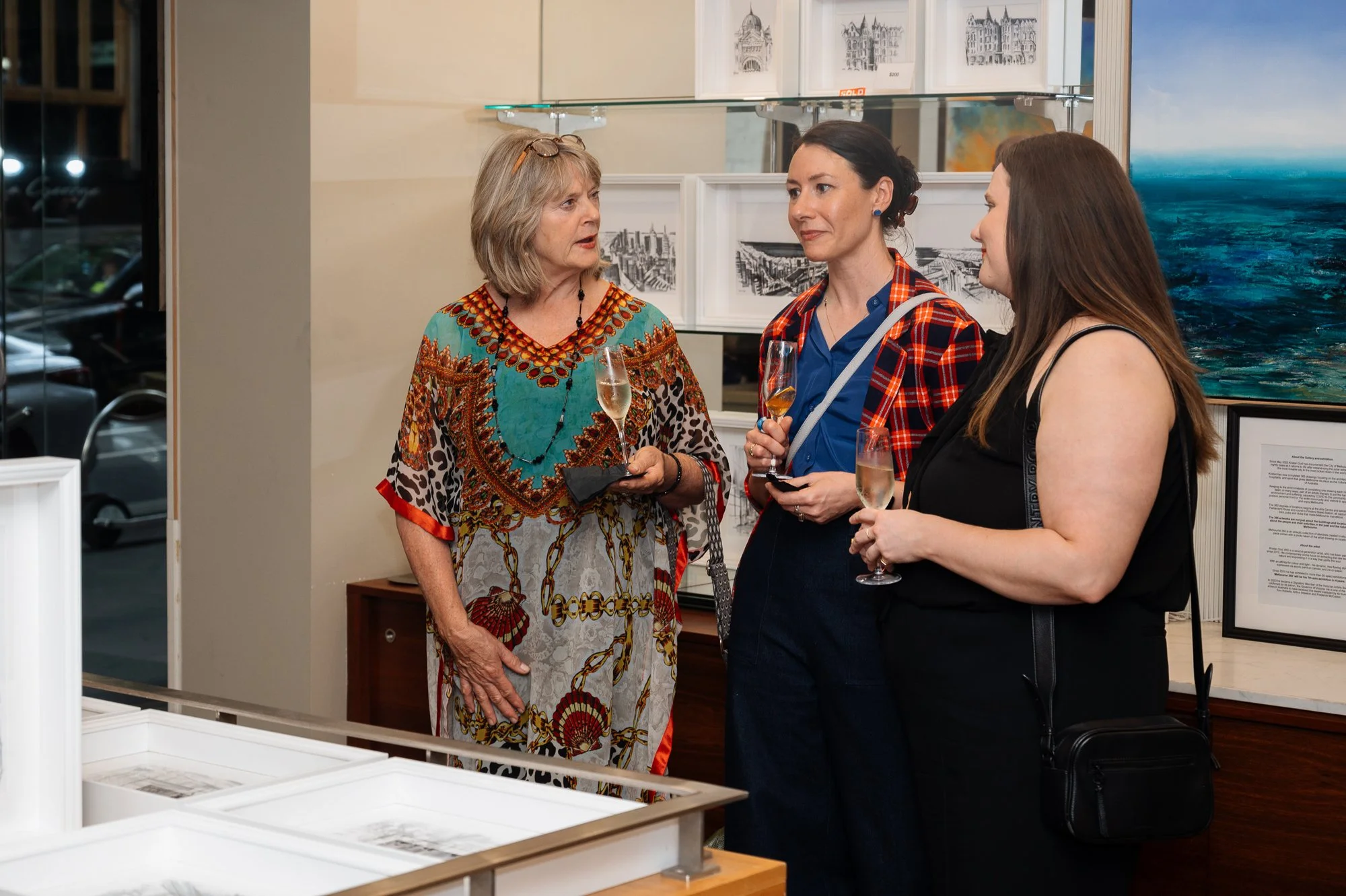 Three women having a conversation at an art gallery, each holding a glass of wine.