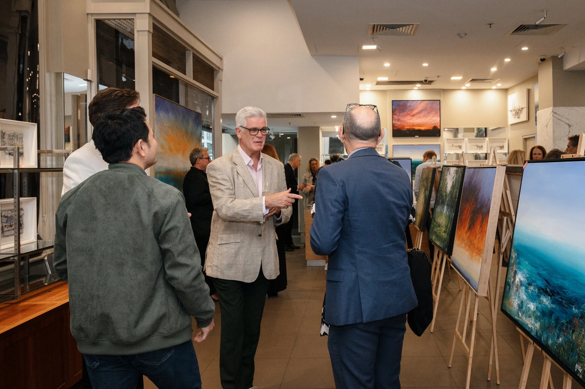 Art gallery with people viewing landscape paintings on display and engaging in conversation.
