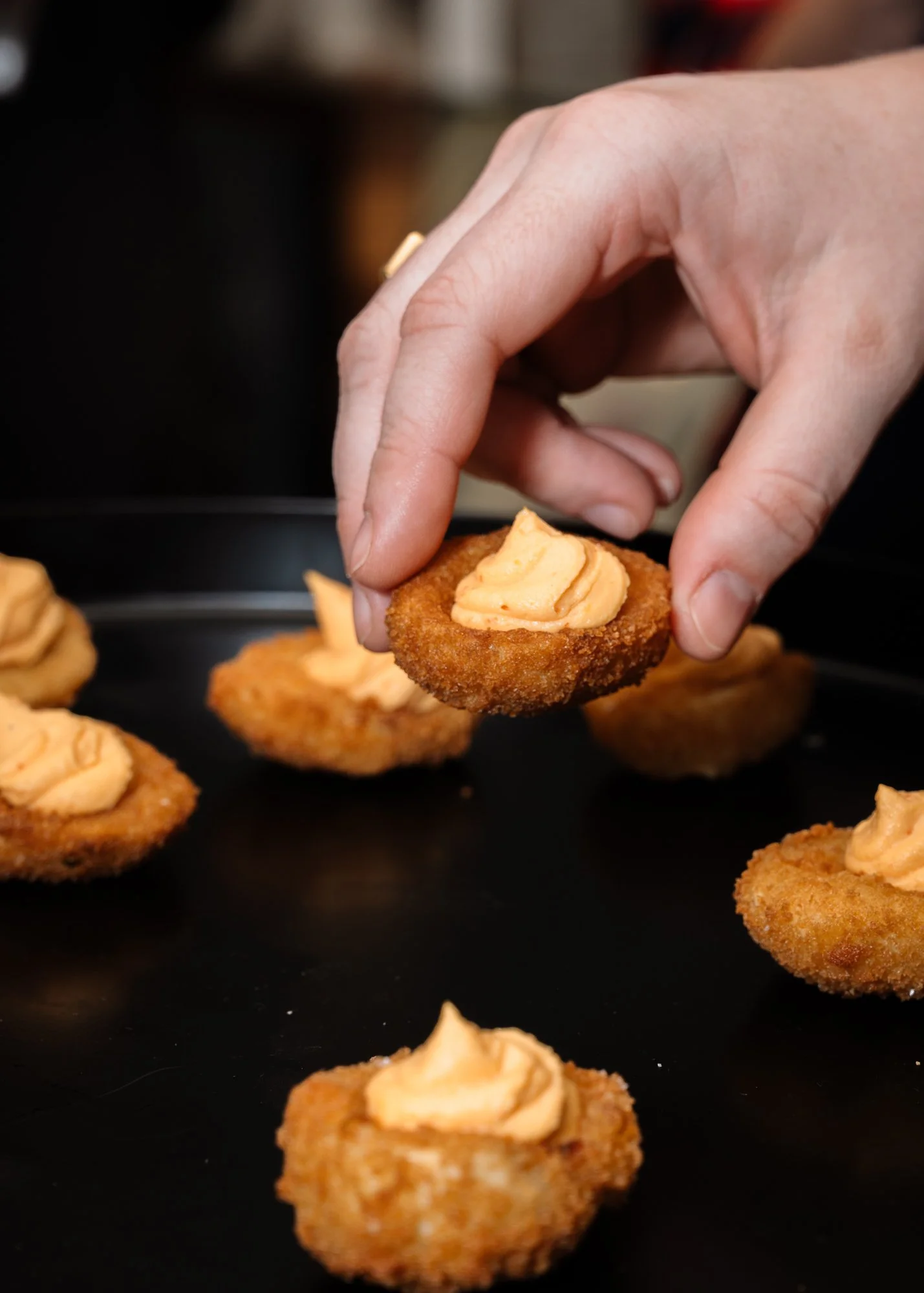 A hand is picking up a mini fried food item with a dollop of orange-colored sauce or spread on top, with more similar items on a dark surface in the background.