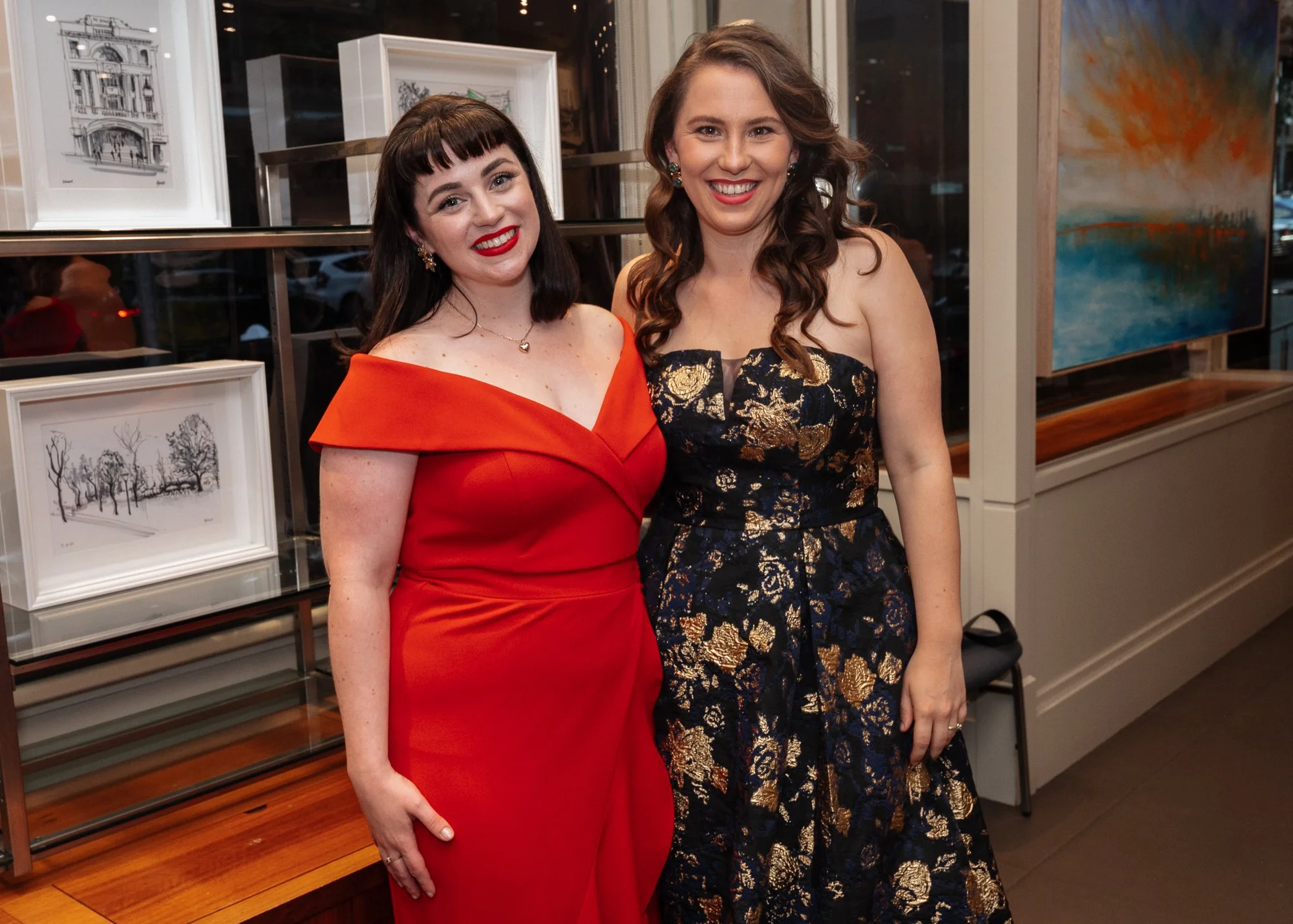 Two women standing side by side, smiling at the camera inside an art gallery. The woman on the left is wearing a red off-the-shoulder dress with dark hair and bangs. The woman on the right is wearing a dark strapless dress with gold floral patterns a