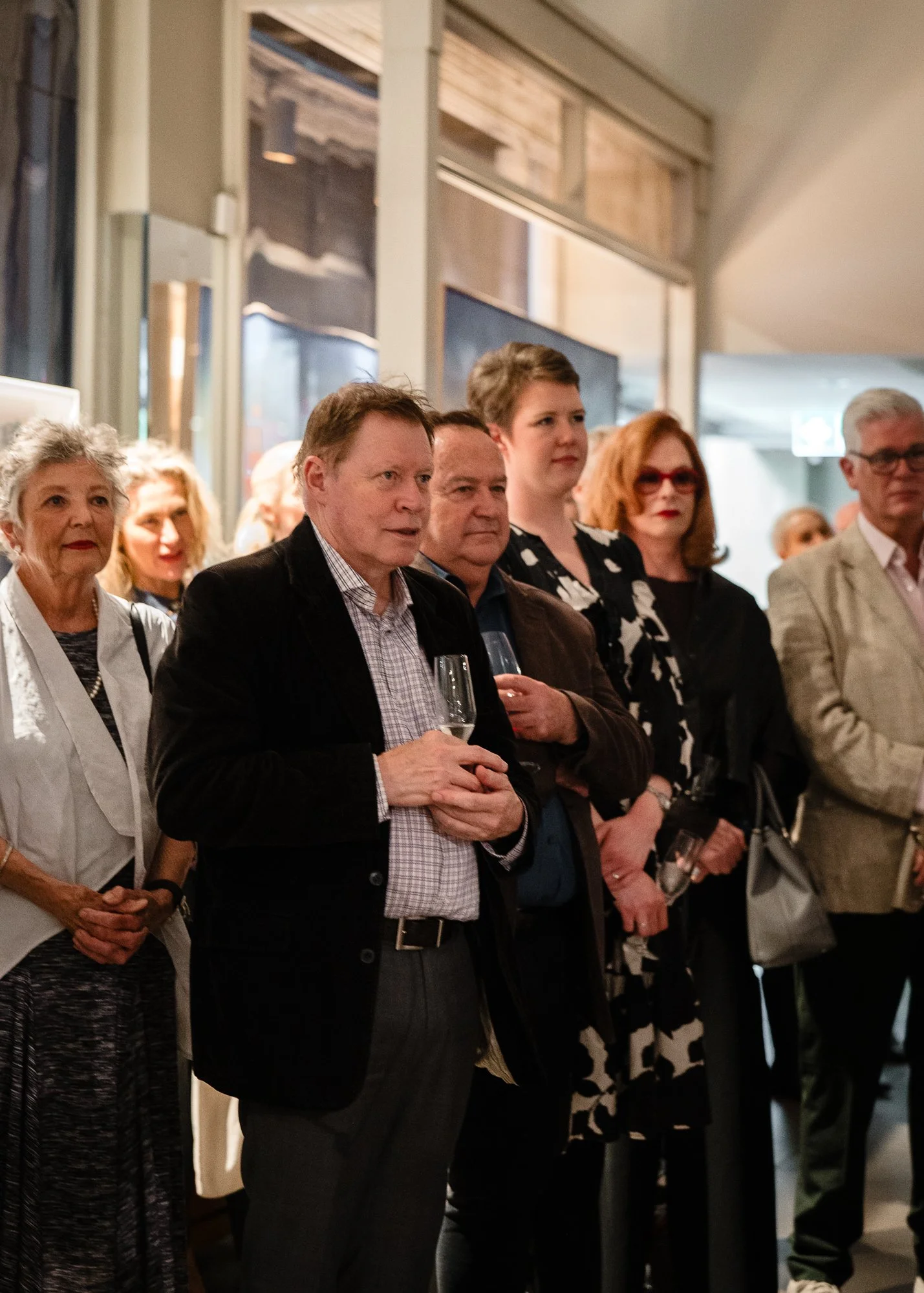 A group of people at an indoor event, some holding glasses of champagne, standing and listening attentively.