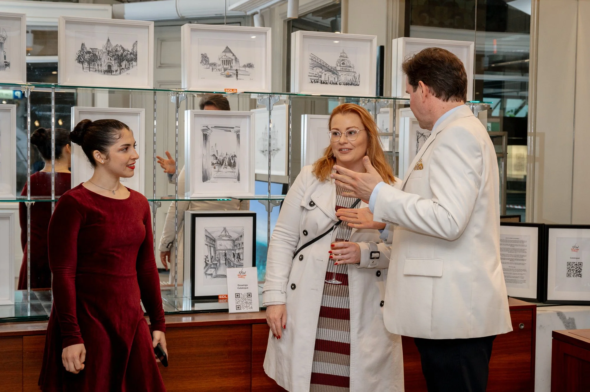 Three people having a conversation at an art exhibit. The woman on the left has dark hair in a bun and is wearing a red dress. The woman in the middle has red hair, glasses, and is wearing a white coat, holding a glass of wine. The man on the right h