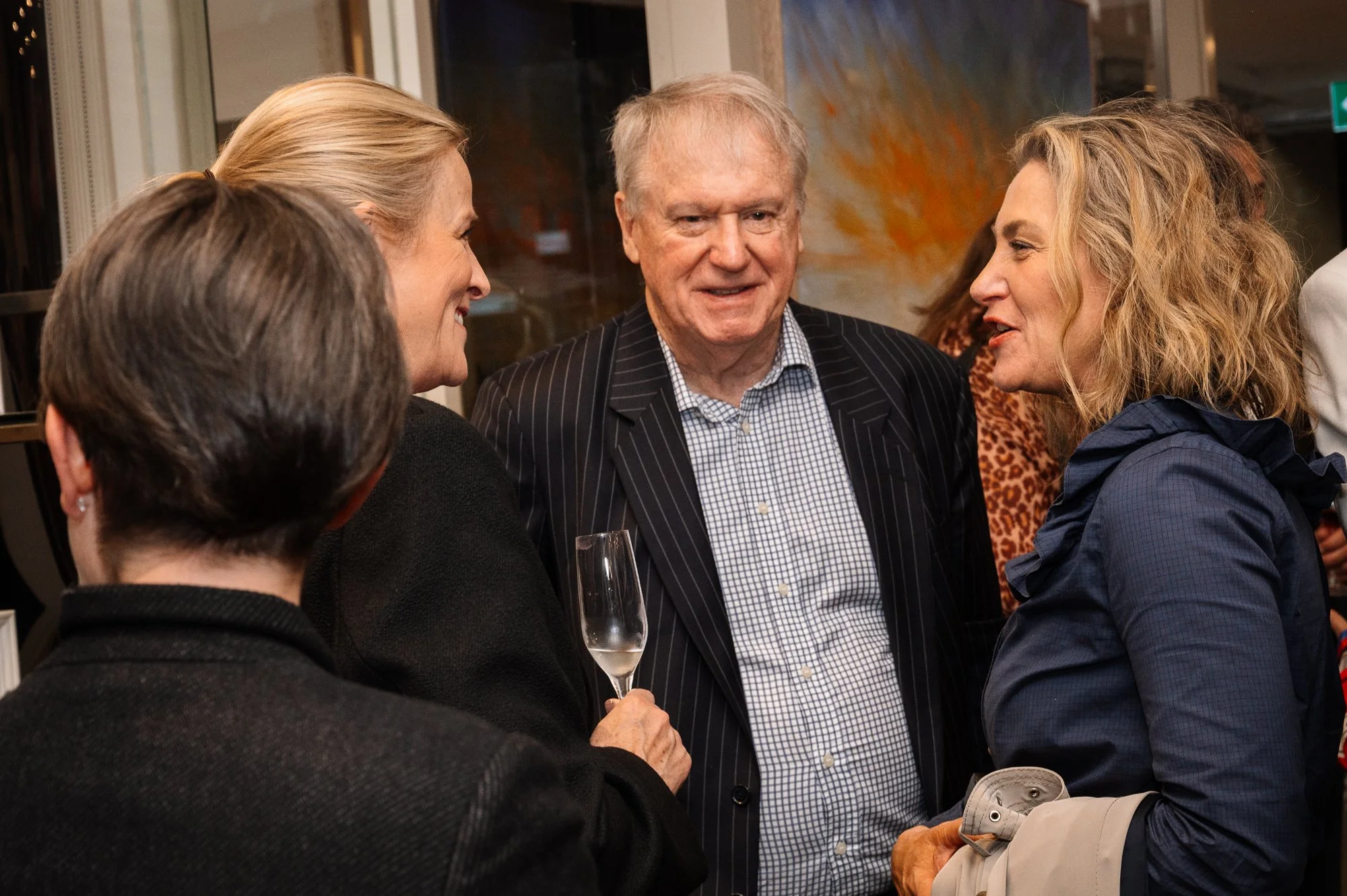 Group of four adults socializing at a gathering, one woman holding a champagne glass, chatting and smiling, in an indoor setting with artwork on the wall in the background.