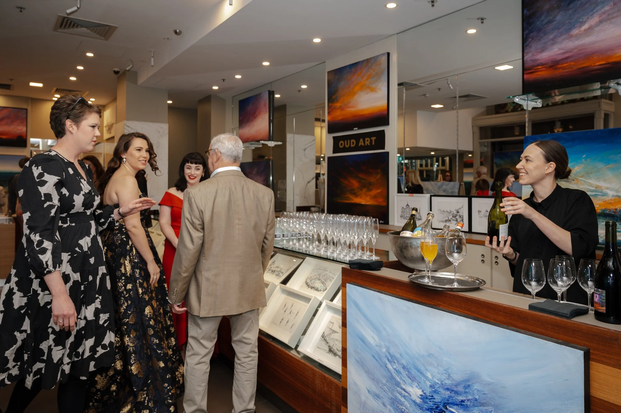 People at an art gallery social event, some are conversing with an older man, while a woman in black serves drinks behind a bar with artwork displayed on walls.