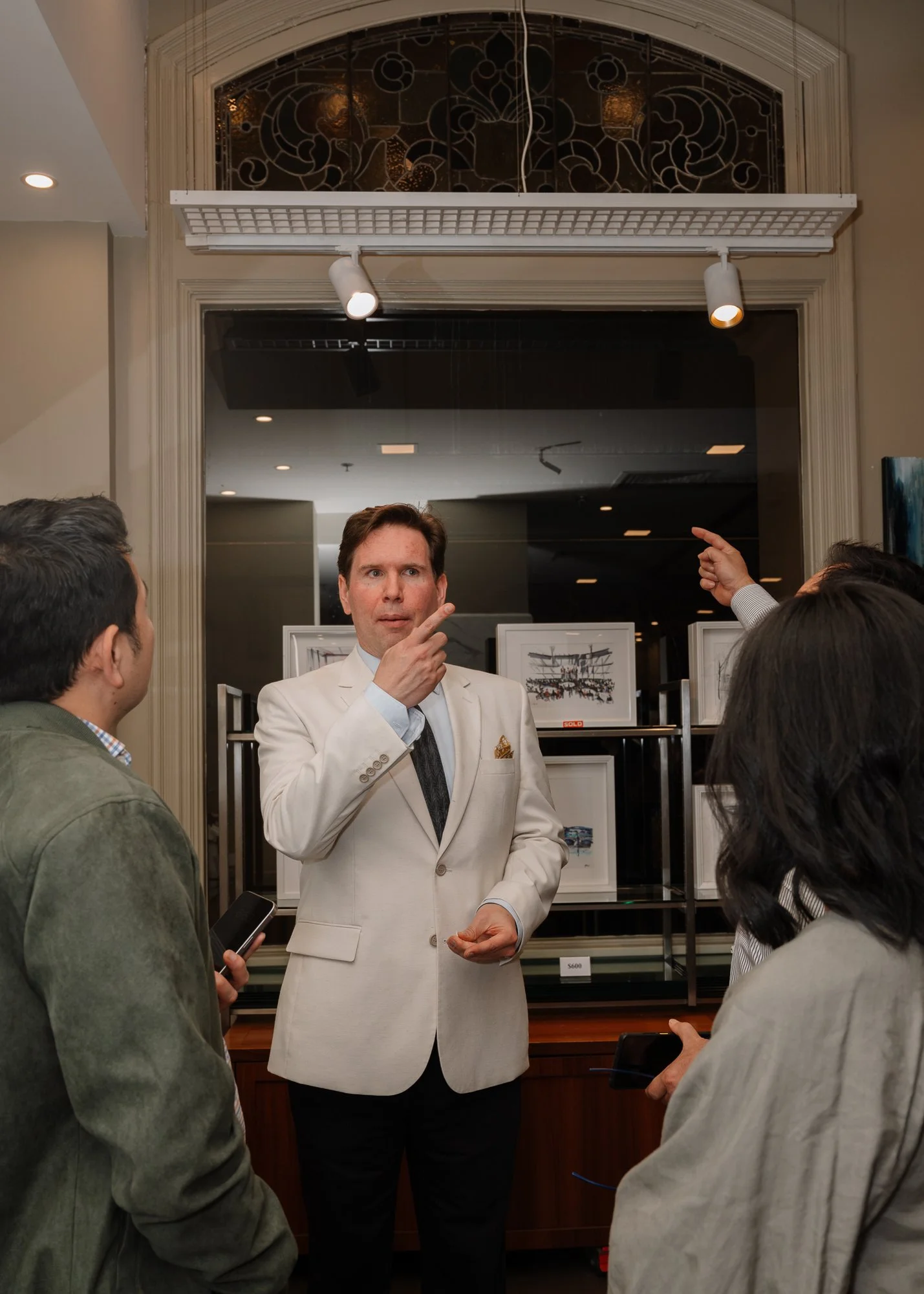A man in a white suit jacket explaining something to a group of people indoors, with artwork displayed on a black rack behind him.