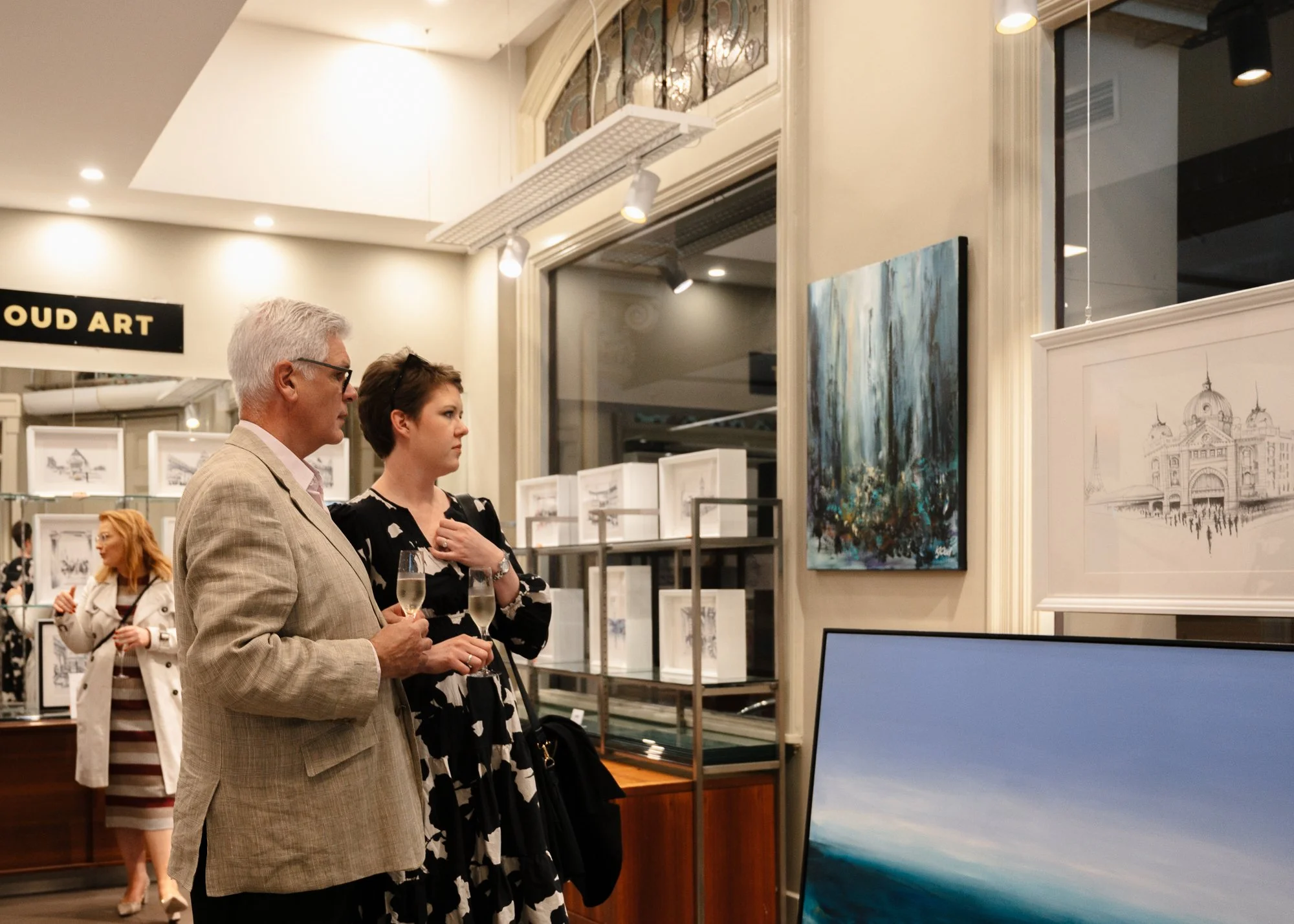 People attending an art gallery, viewing paintings, with two individuals holding champagne glasses in the foreground. Art pieces and display shelves are visible in the background.