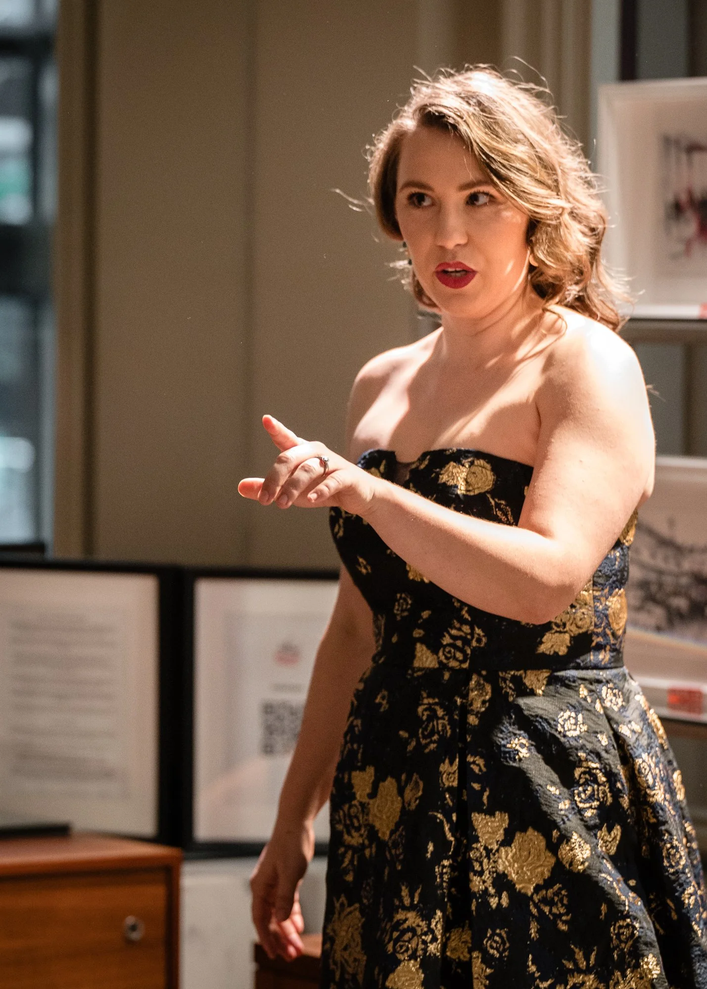 A woman with shoulder-length wavy hair, red lipstick, wearing a strapless black dress with gold floral patterns, gesturing with her right hand while talking indoors.