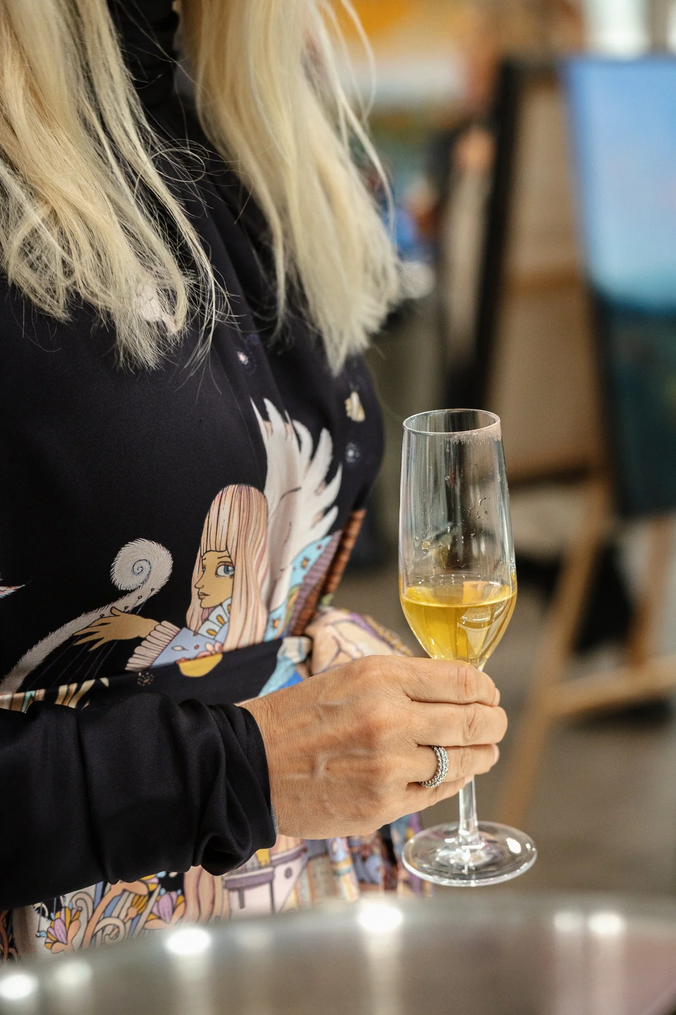 A woman with blonde hair wearing a colorful, patterned shirt is holding a flute of white wine or champagne in a social setting.