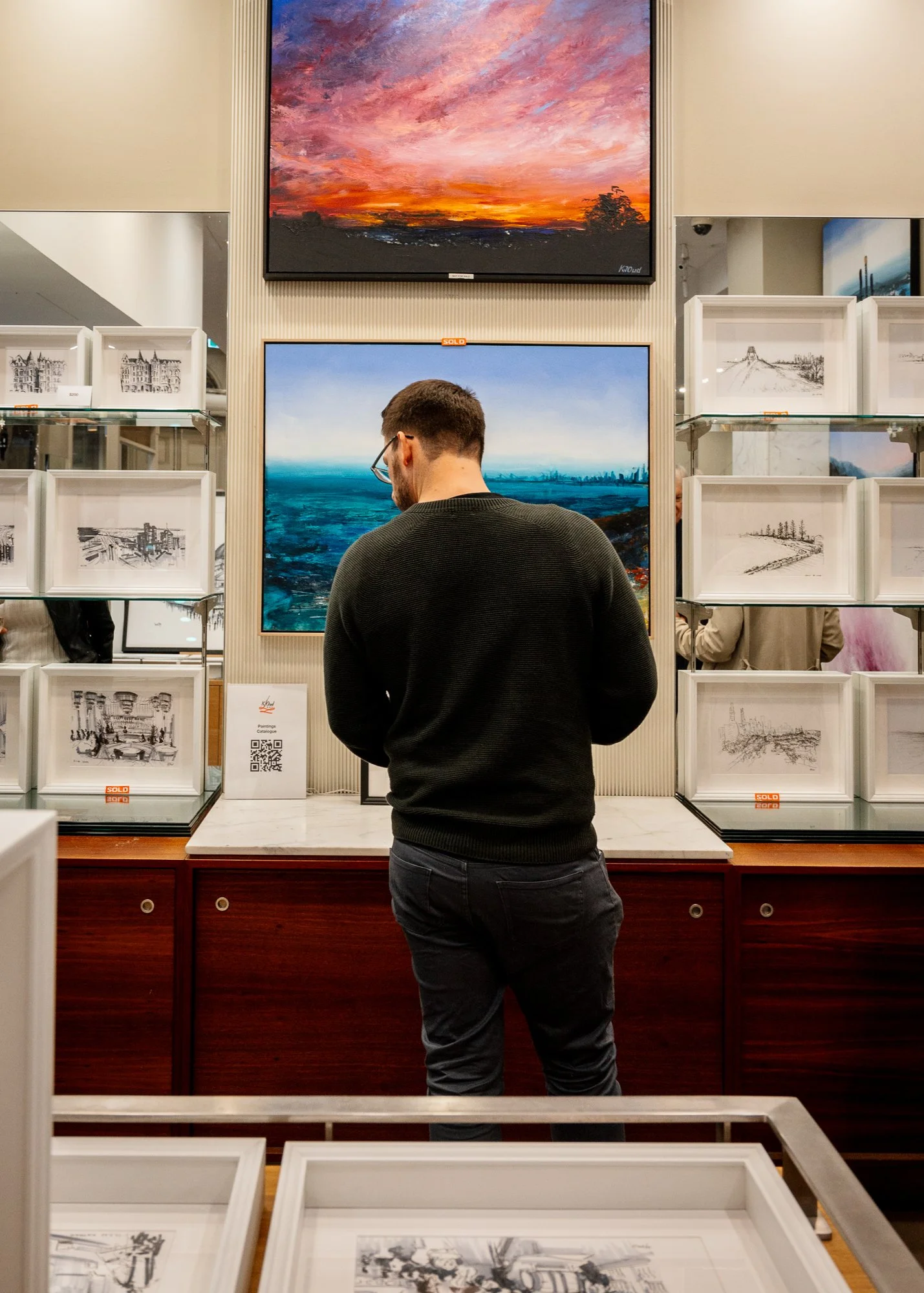 A person with glasses wearing a black sweater and dark pants looking at a painting in an art gallery. Two large landscape paintings are mounted on the wall behind them. There are framed sketches in glass display cases on either side of them, with som