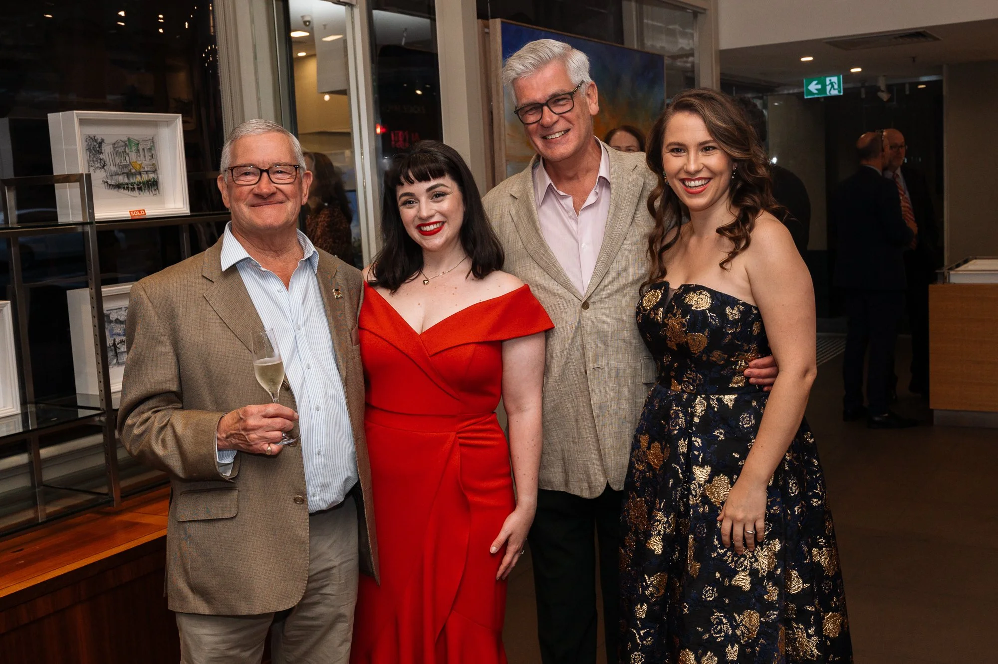 A group of four people dressed in formal attire at an indoor event, smiling for a photo. They are standing close together, with a backdrop of artwork and people in the background.