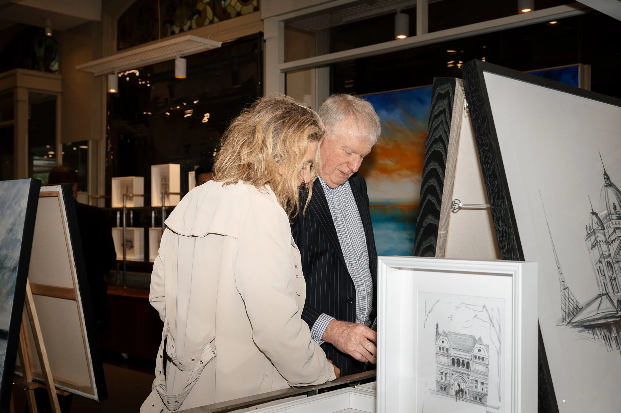 Two people looking at artwork in an art gallery. The artwork includes sketches and paintings, with some framed and displayed on easels.