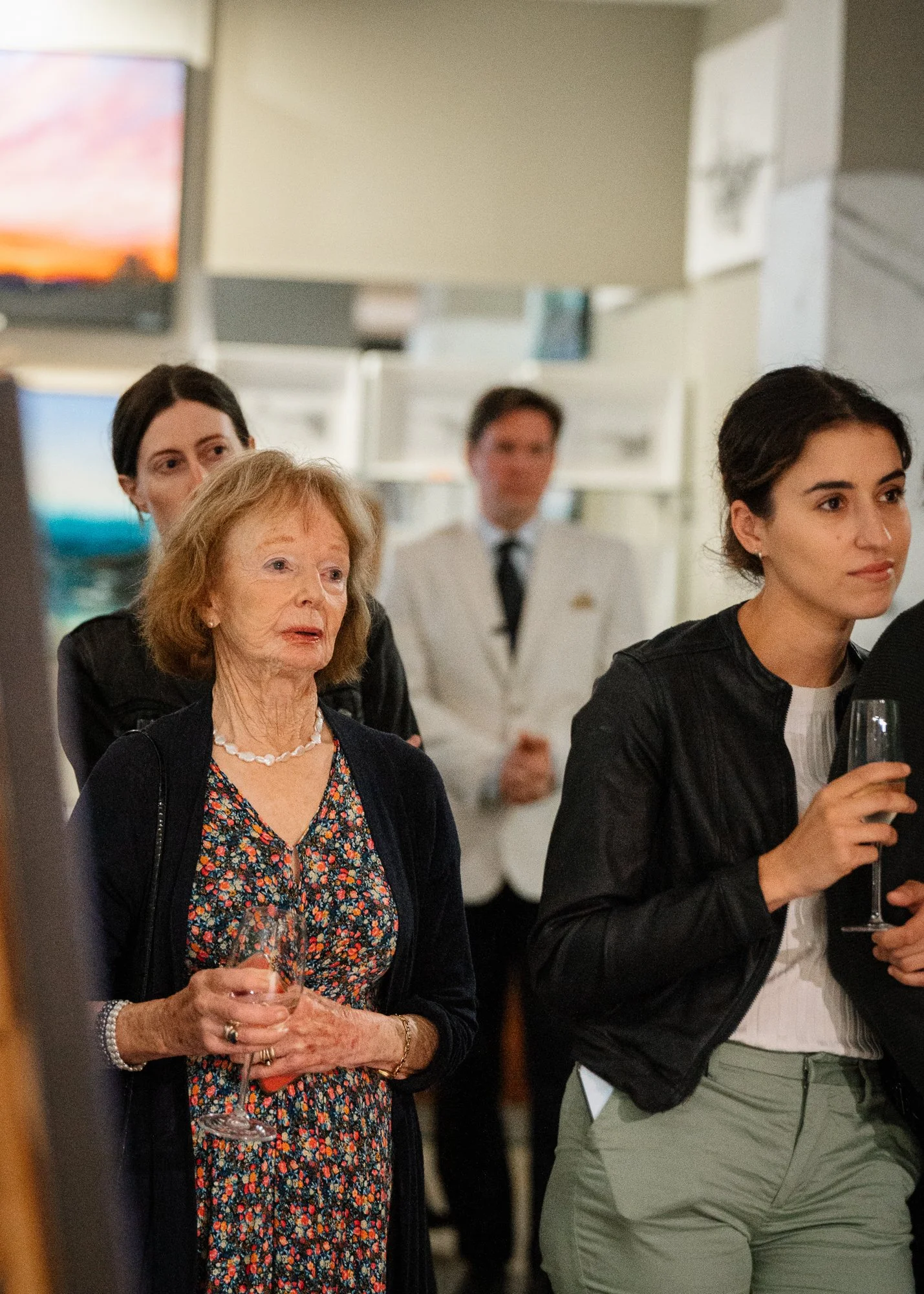 Group of four people, including an older woman with short curly hair, holding a wine glass, at what appears to be a social gathering or event.