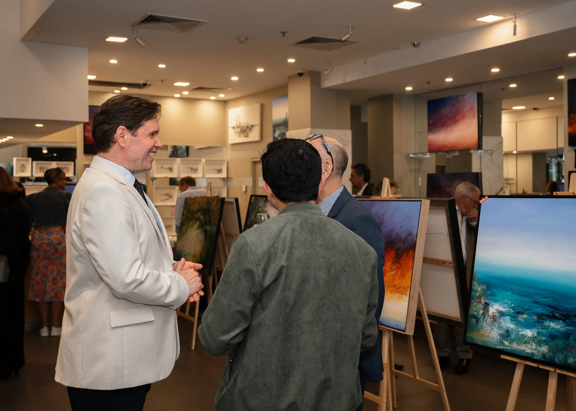 Three men engaged in conversation at an art gallery, surrounded by various landscape paintings on easels.