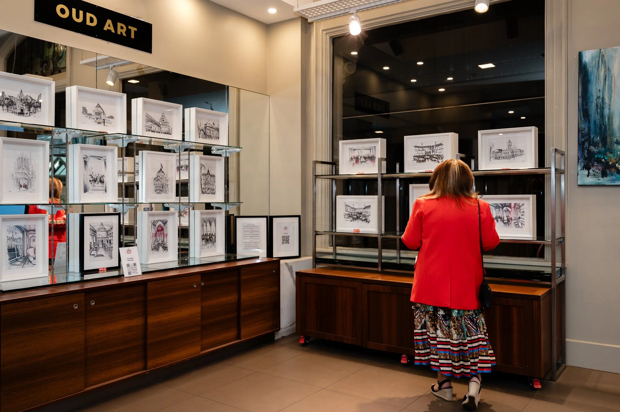 A woman in a red coat and patterned dress is viewing framed artwork and sketches displayed on metal and glass shelves inside an art gallery. The sign above reads 'Oud Art'.