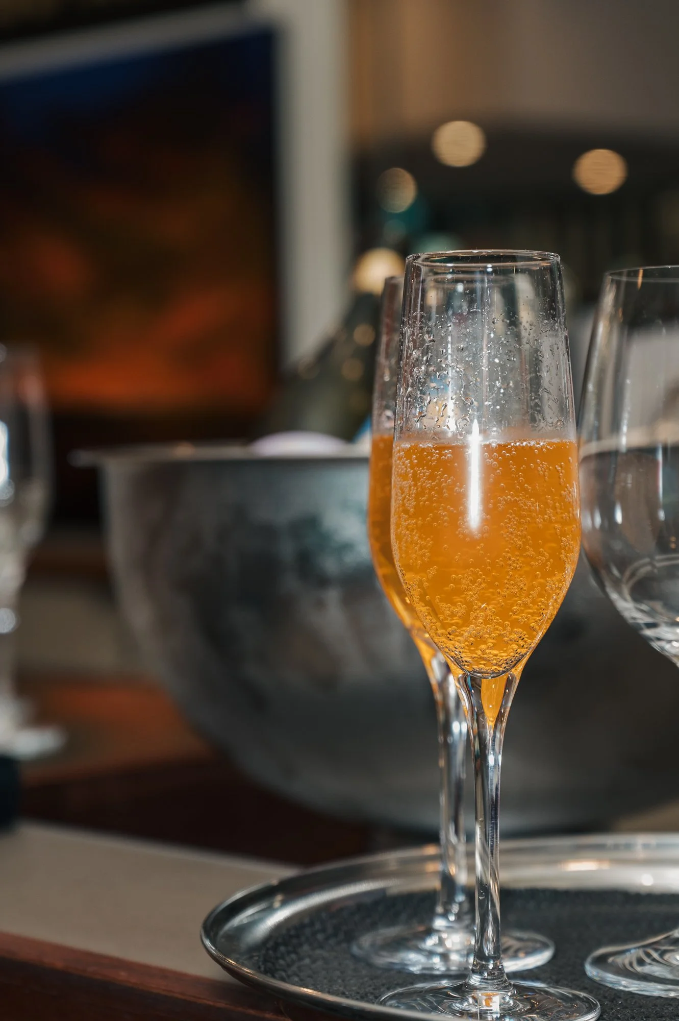 A glass of sparkling champagne or sparkling wine on a table with other glasses and a silver bucket in the background.