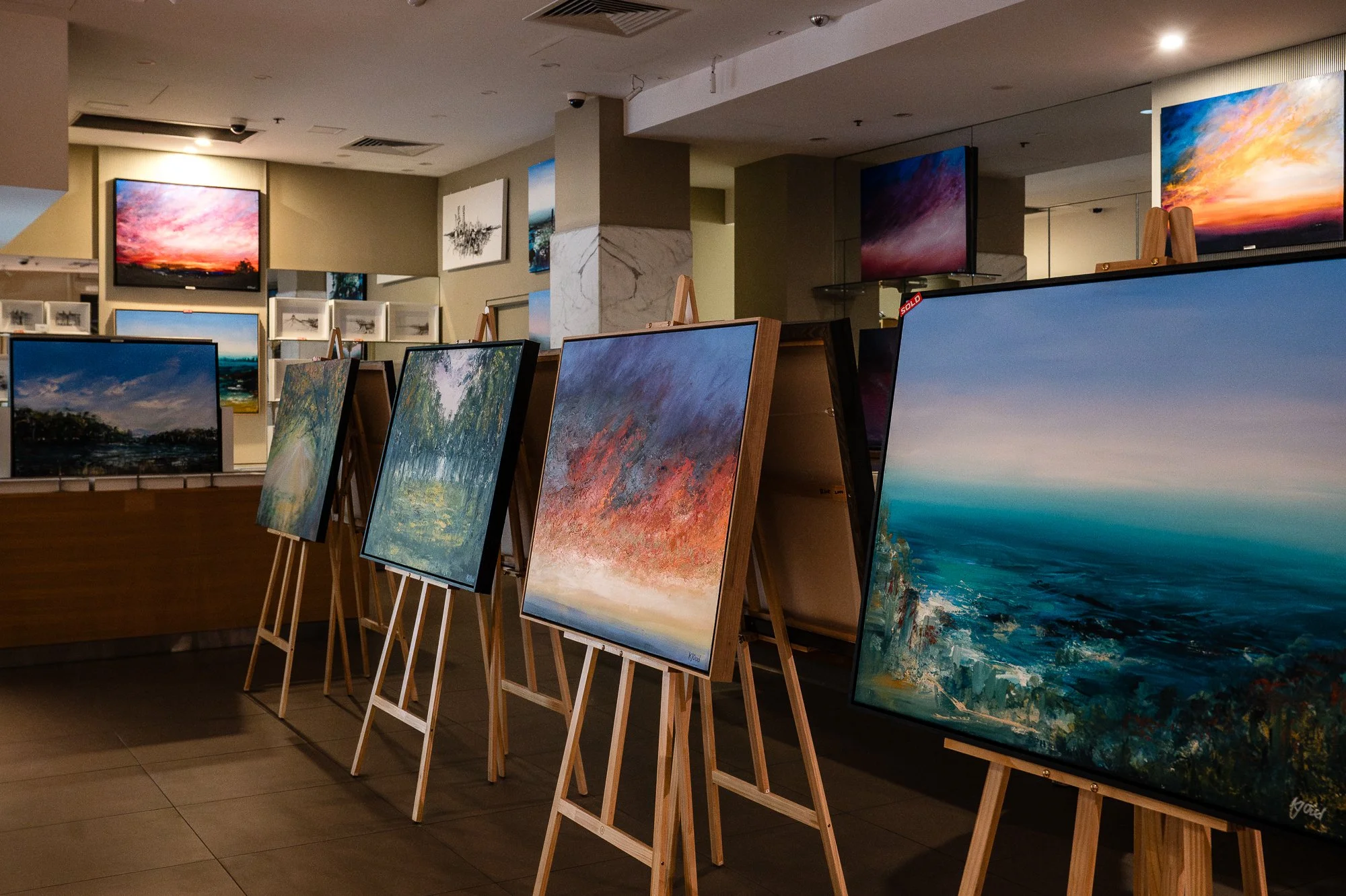 An art gallery displays colorful landscape paintings on wooden easels, with more paintings hung on the walls in the background.