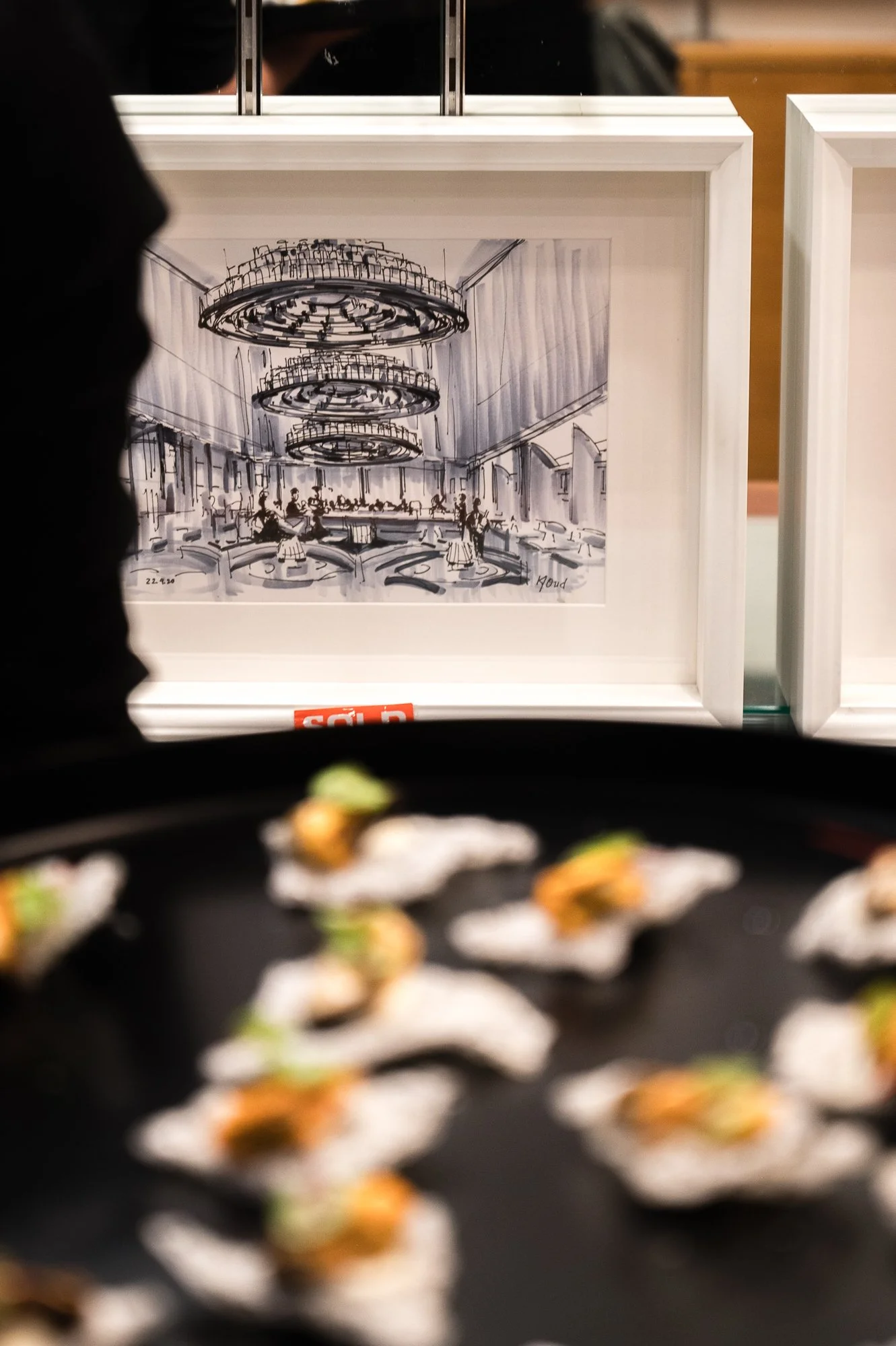 A framed sketch of an elegant hotel lobby with chandeliers and people dining, displayed behind a tray of food served on small plates.