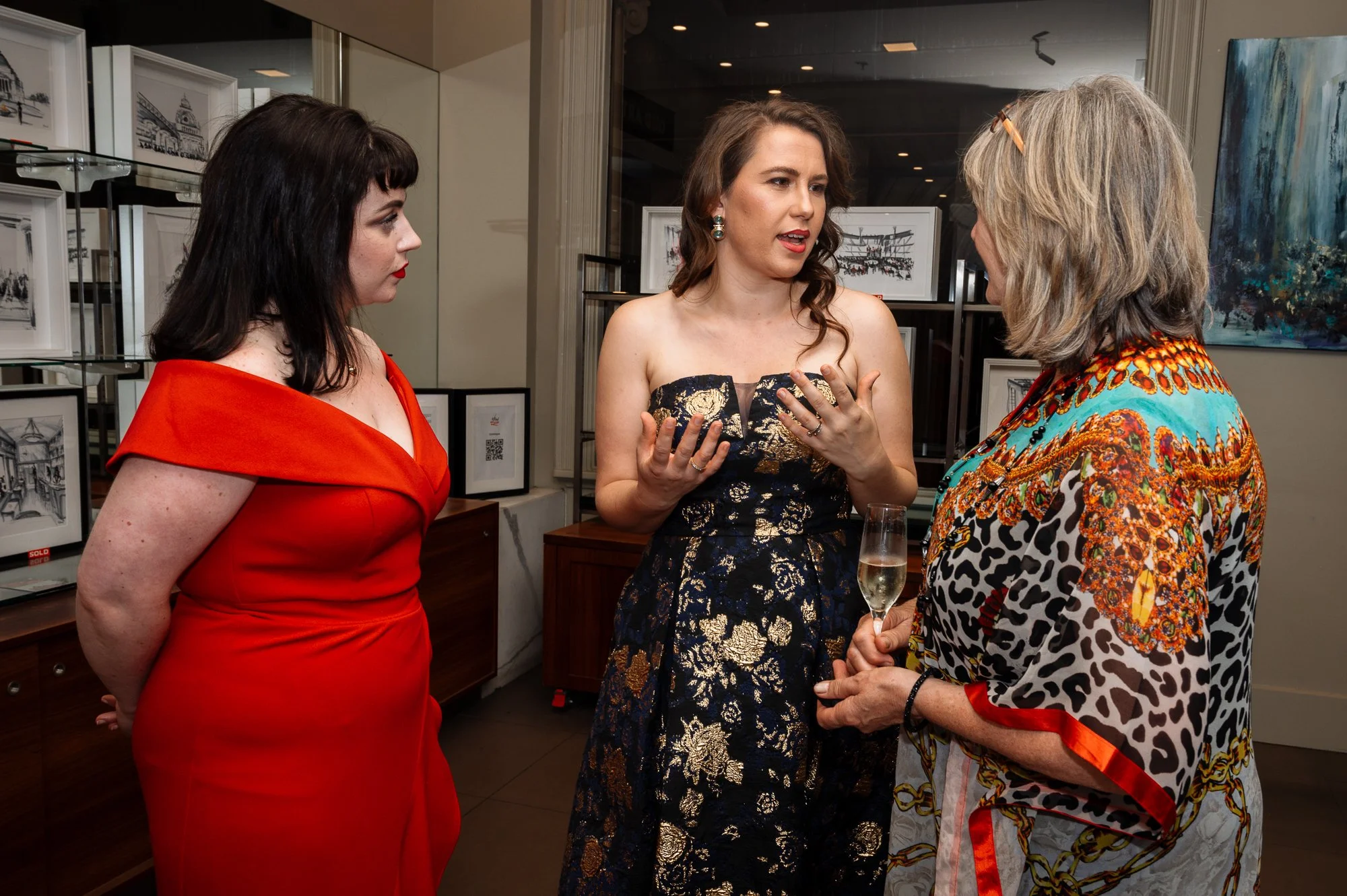 Three women engaged in conversation at an art gallery or exhibition, one holding a champagne glass, with framed artwork on display in the background.
