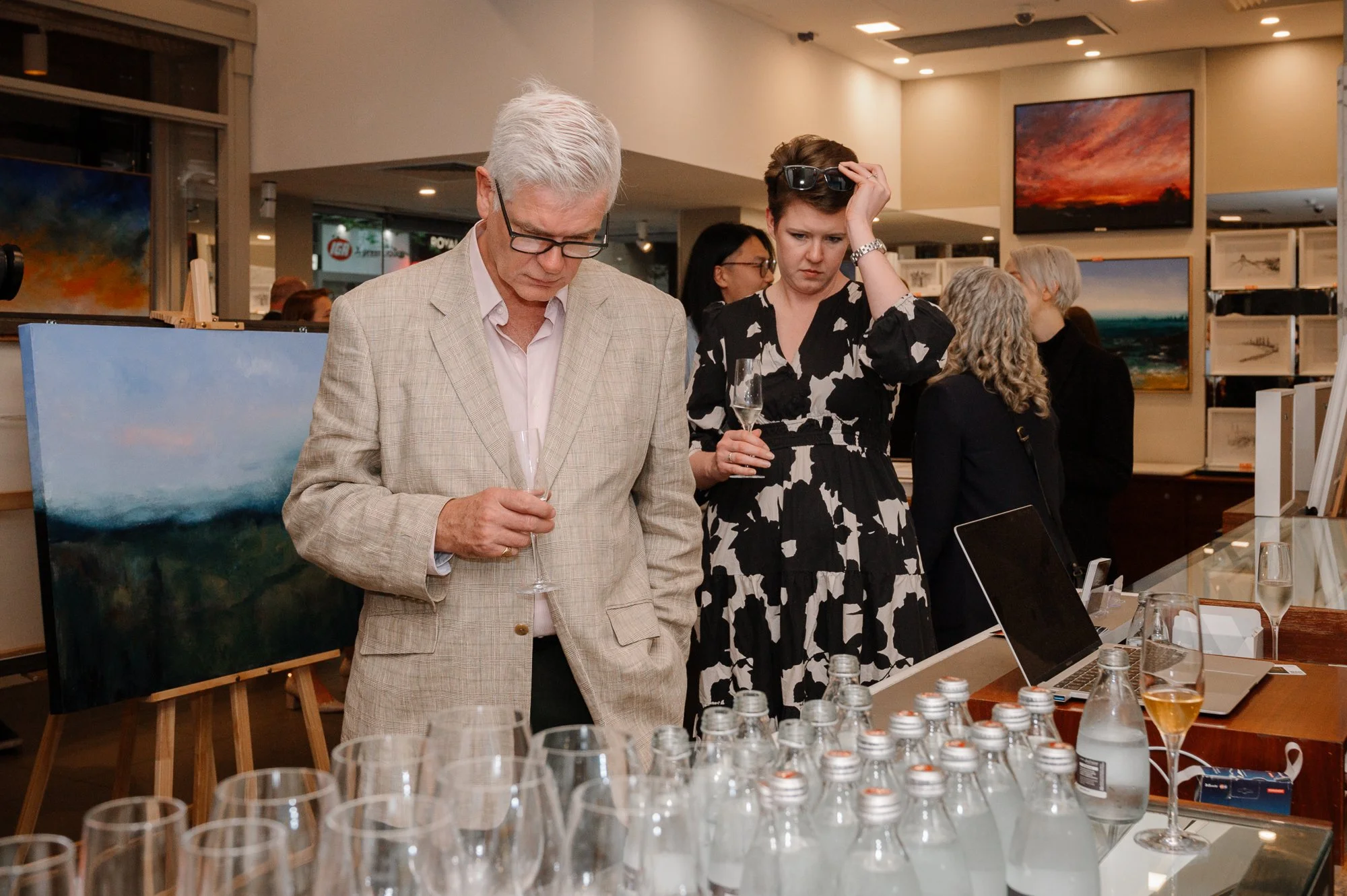 Two people at an art gallery event, an older man in a beige suit and a woman in a black and white dress, standing by a table with glasses of sparkling beverage and bottles, with paintings displayed on the walls behind them.