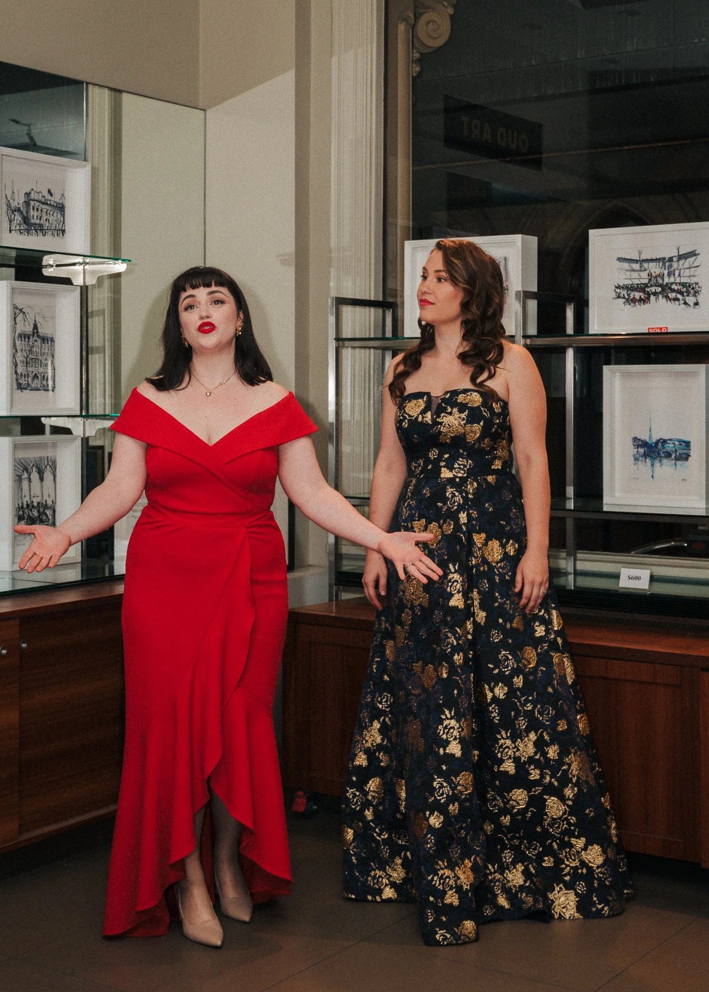 Two women in elegant dresses standing and speaking indoors, with artwork displayed in the background.