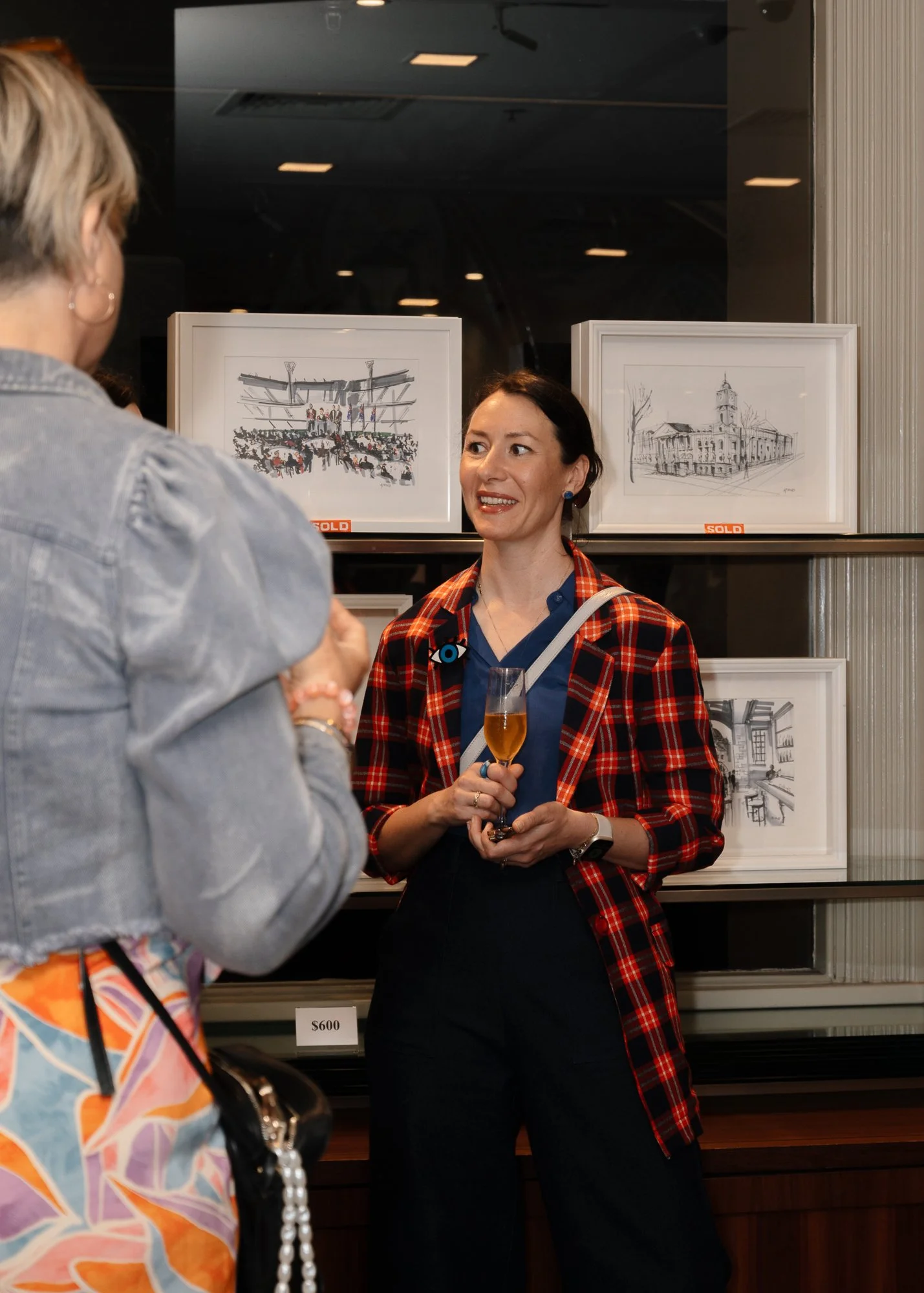 Two women are talking at an art gallery or exhibit. One woman is wearing a gray jacket with colorful pants, and the other woman is holding a glass of wine and wearing a red plaid blazer with a blue shirt underneath.