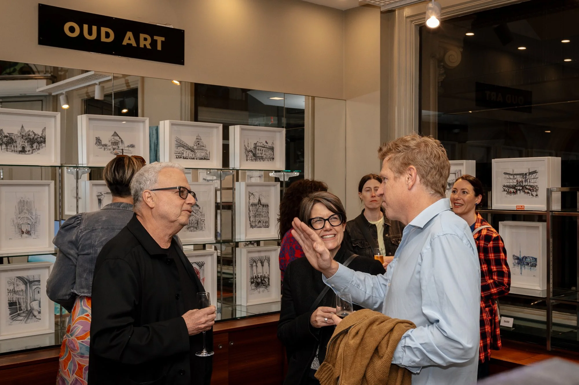 People attending an art gallery event, engaged in conversation, with framed artwork displayed on shelves behind them.